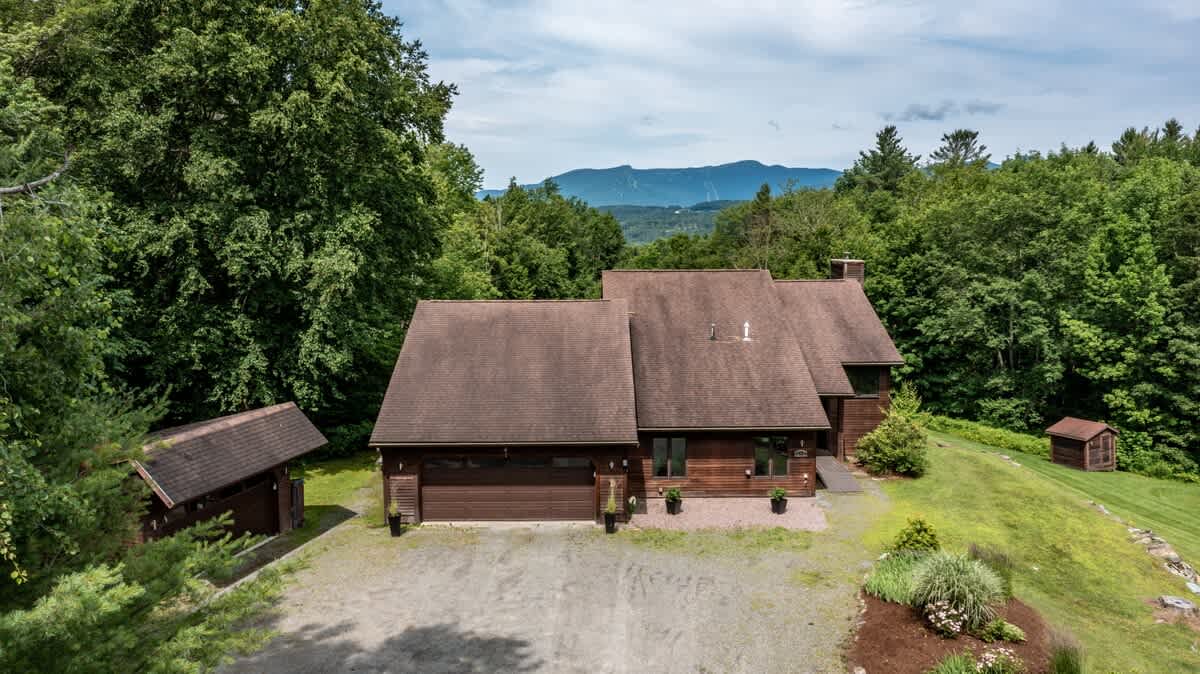 Aerial view of the front of a large brown house with a two-car garage, surrounded by green lawns and dense trees, with distant mountains visible under a partly cloudy sky.