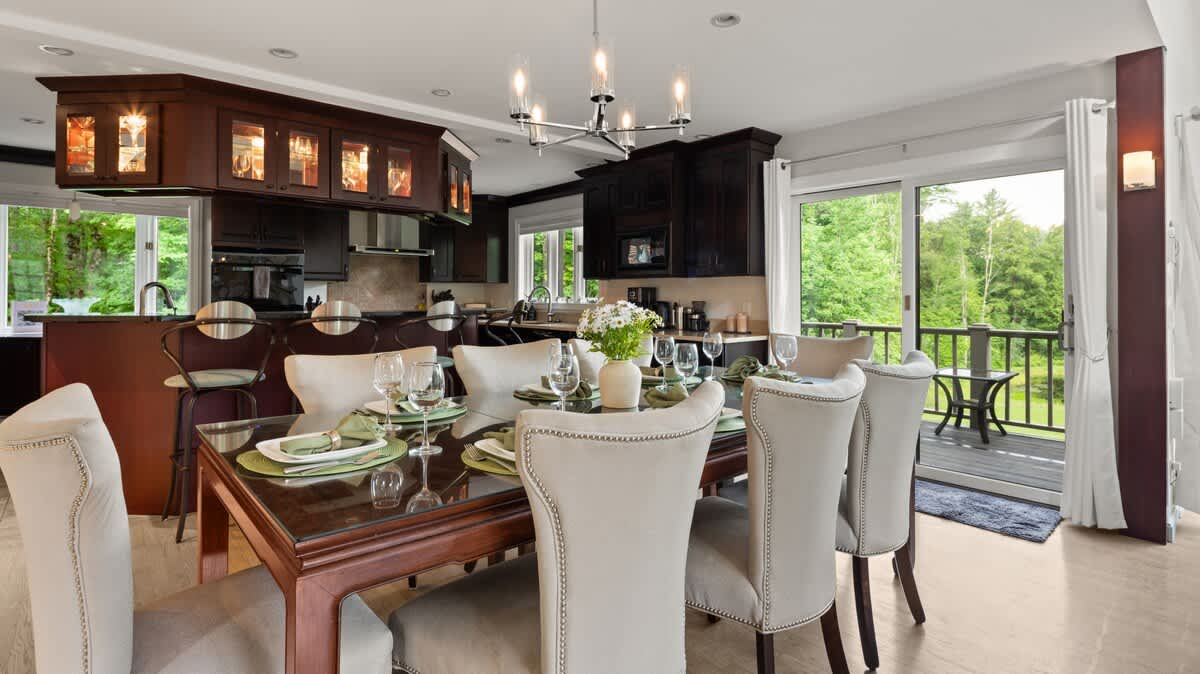 An elegant dining area features a dark wood table set for six with upholstered chairs, adjacent to a modern kitchen with dark cabinetry and a breakfast bar, and offering access to an outdoor deck through sliding glass doors.