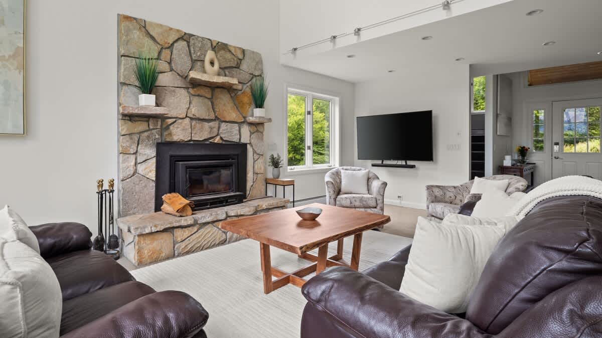 A cozy living room features a prominent stone fireplace with a wooden mantel, dark leather sofas, and patterned armchairs arranged around a rustic wooden coffee table, with large windows offering views of the surrounding greenery.