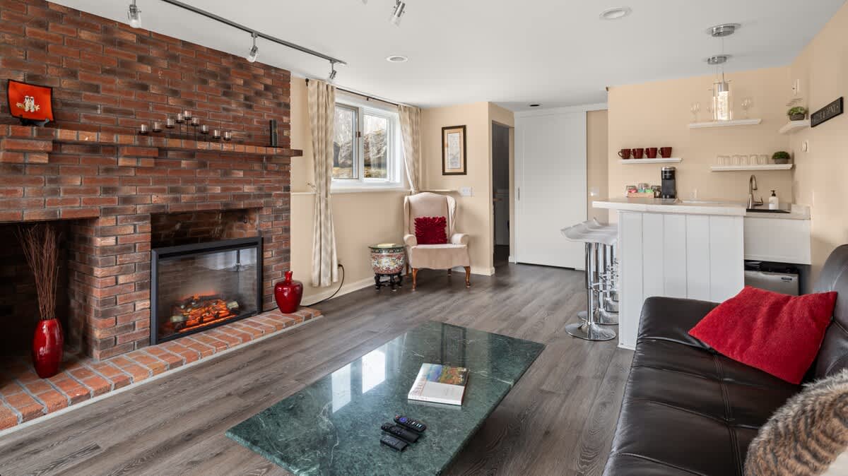 A cozy living room featuring a prominent brick fireplace with a modern electric insert, a dark green marble-top coffee table, and a wet bar with white cabinetry and bar stools. The room has dark wood-look flooring.