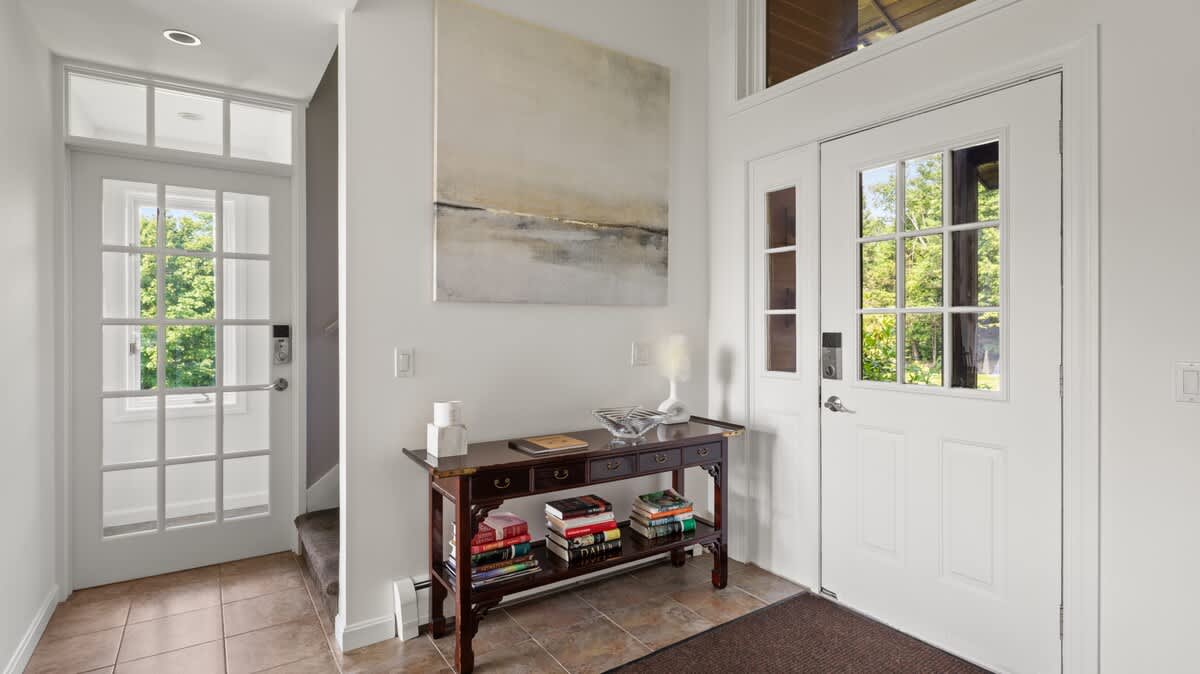 Inviting entryway with light-colored tiled floors and white walls, featuring a dark wood console table adorned with books and decor. Two white doors with glass panes, one leading outside and another to an interior space, allow natural light to fill the area.