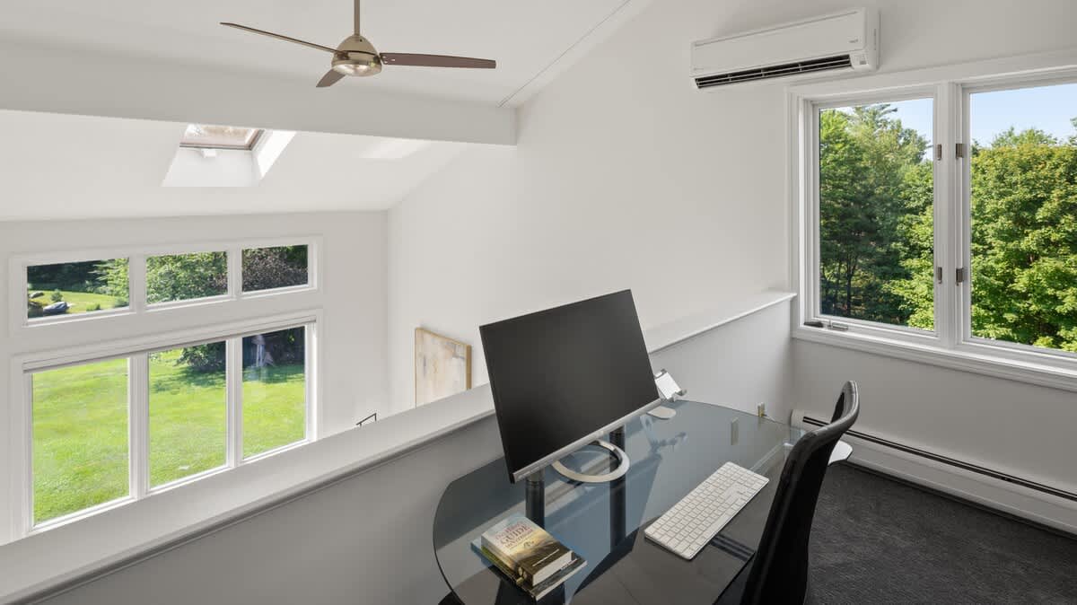 Bright loft workspace with a glass desk and black office chair, overlooking a lower level and offering expansive views of a green lawn and trees through large windows. A ceiling fan and a mini-split AC unit are visible, along with skylights.
