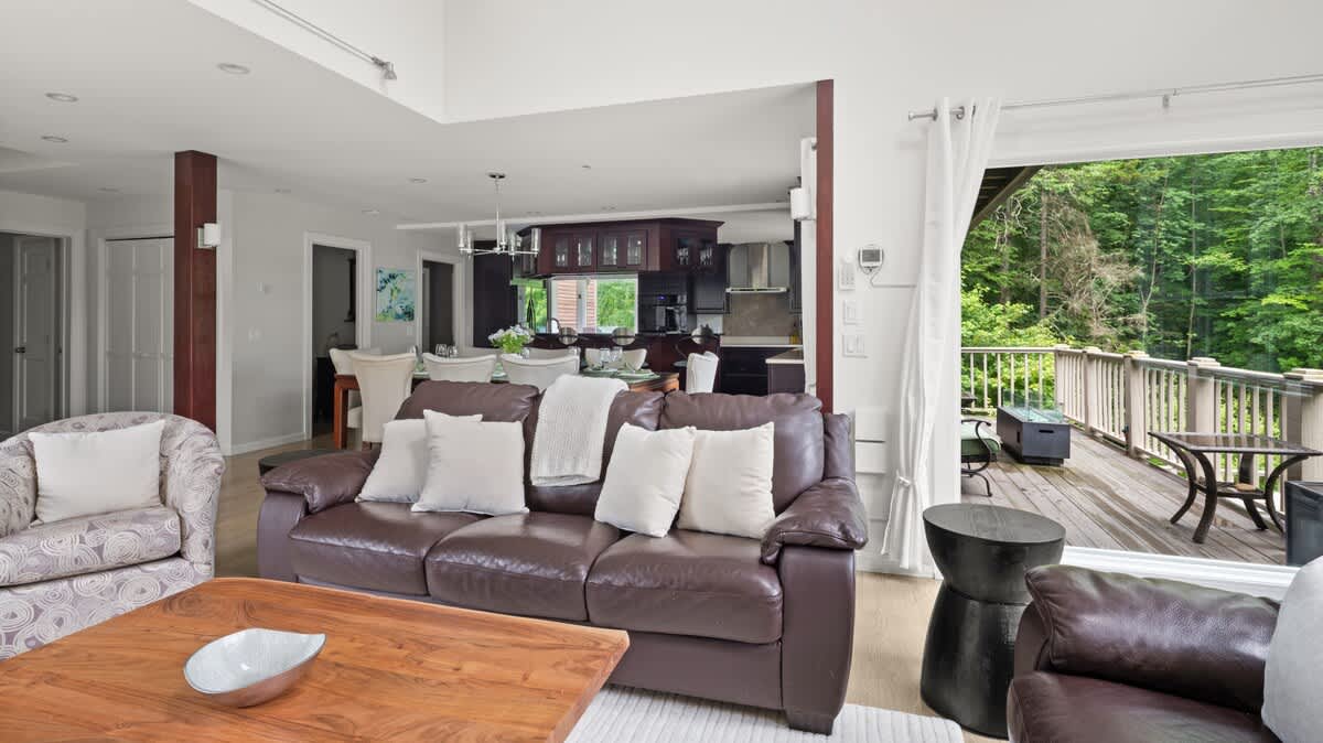 An open-concept living space with dark leather sofas and a view towards the kitchen and dining area, featuring a large sliding glass door that opens to an outdoor deck surrounded by trees.