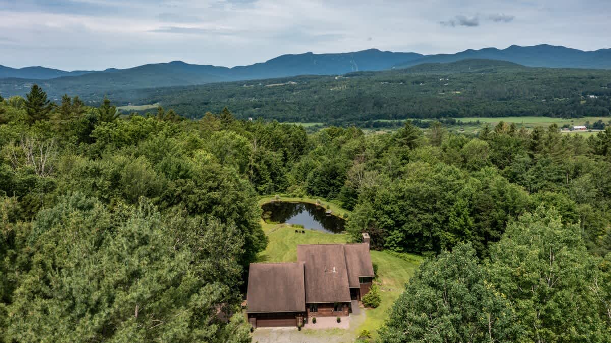 Aerial view of a large brown house nestled among lush green trees, with a small pond reflecting the sky and surrounding foliage, and distant mountains under a cloudy sky.