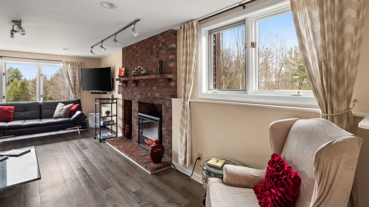 A spacious living room with a black futon sofa, a large brick fireplace with an electric insert, and a window overlooking a wooded area. A flat-screen TV is mounted on the wall.