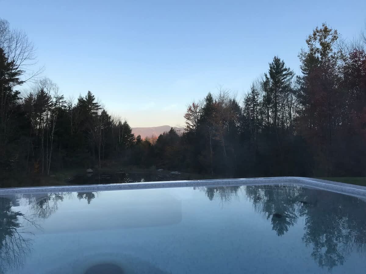 View from inside a hot tub looking out over a tranquil pond and dense forest, with distant mountains under a soft evening sky, highlighting the serene outdoor setting.