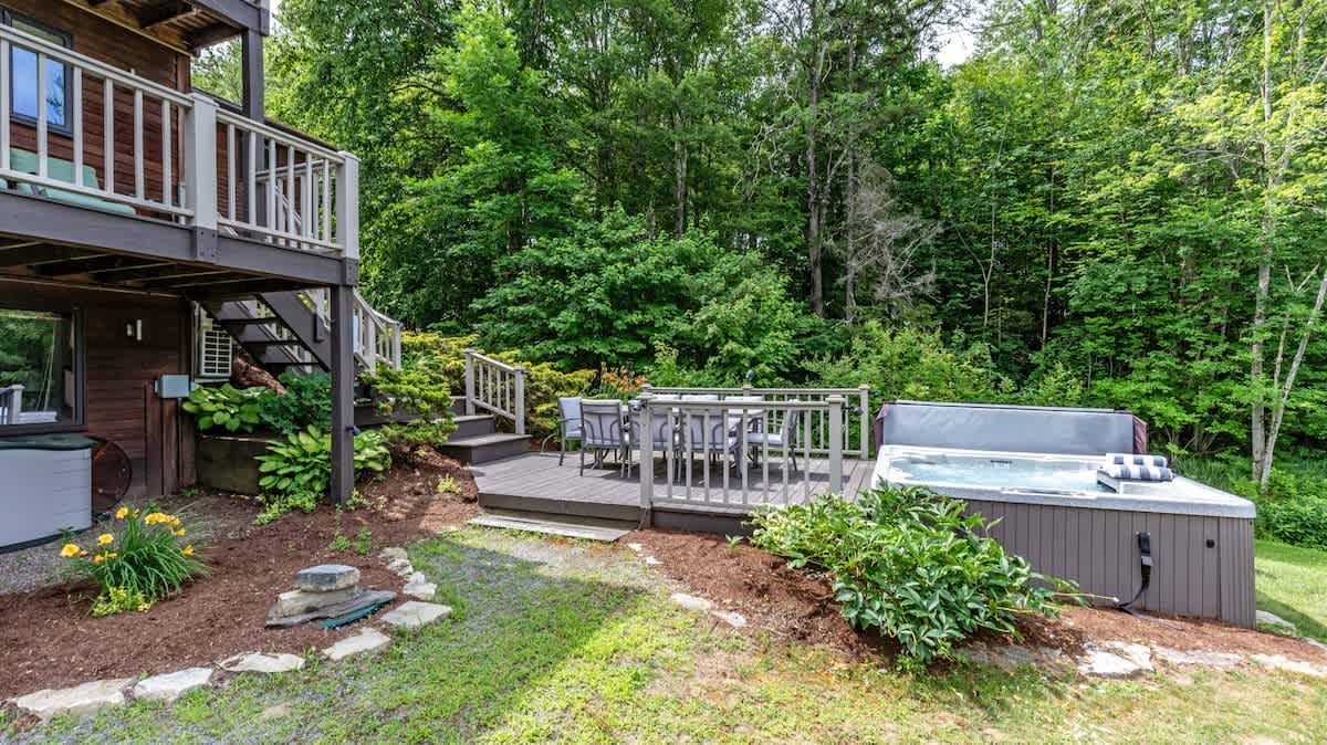 Wooden deck area featuring a hot tub and an outdoor dining set with chairs, nestled beside the house. Steps lead up to a higher level, and the area is surrounded by lush green landscaping and trees.