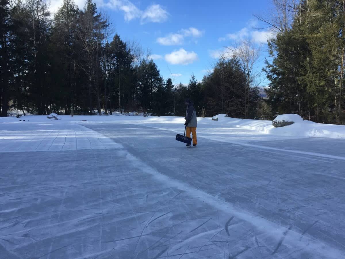 A person skates on a large, frozen outdoor ice rink in a snowy, wooded setting, with bare trees and evergreens under a bright blue sky with scattered clouds.