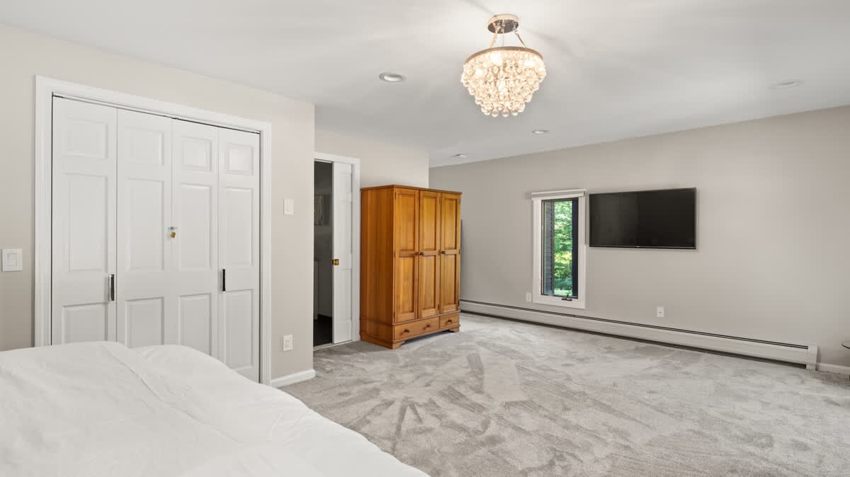 Spacious bedroom with light gray walls and plush gray carpet, featuring a large white bed and a wooden armoire. A wall-mounted TV is visible, and a sparkling chandelier hangs from the ceiling, adding a touch of elegance.