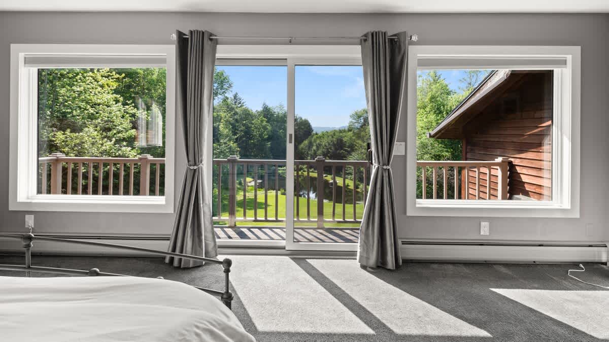 A bright bedroom featuring a large sliding glass door and two side windows, offering panoramic views of a wooden deck, a serene lake, and lush green trees. The room has light gray walls and carpet, with dark gray curtains framing the view.