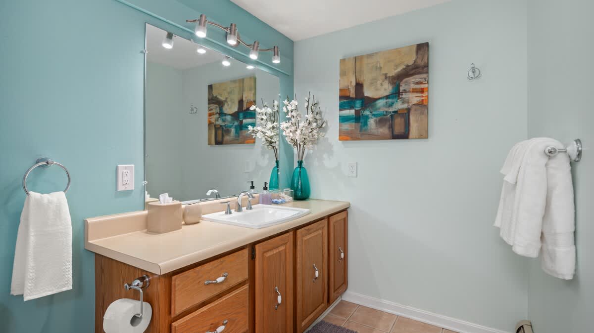 A bathroom with light blue walls, a wooden vanity with a single sink, a large mirror, and modern light fixtures. Abstract artwork hangs on the wall.