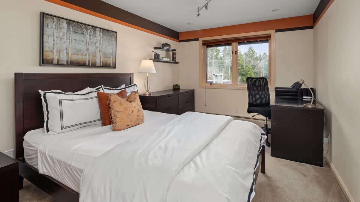 A bedroom with a dark wood bed frame, white linens, and a desk area with an office chair by a window. The room has light-colored walls with a dark brown and orange accent border.
