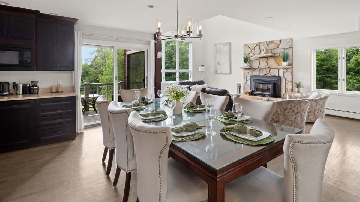 Elegant dining area with a large rectangular table set for eight, featuring upholstered chairs and a modern chandelier. It is open to the living room with a stone fireplace and offers access to an outdoor balcony.