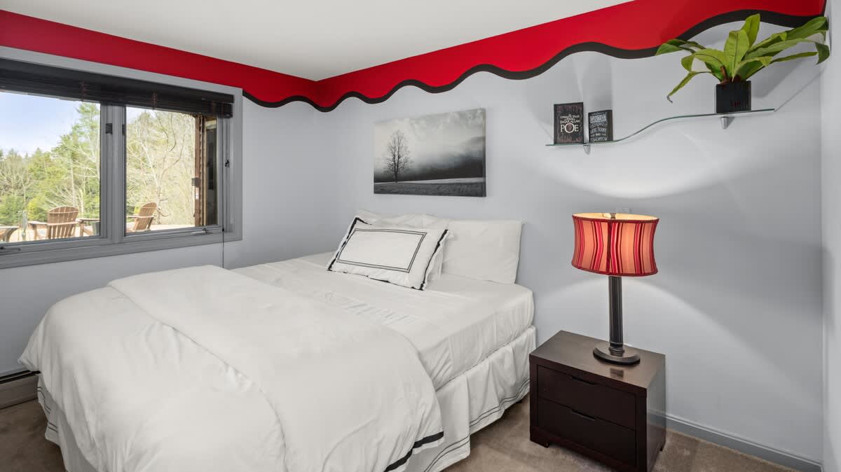 A bedroom with light blue walls and a distinctive red and black wavy border near the ceiling. It features a white-linened bed, a dark nightstand with a red lamp, and a window with outdoor views.