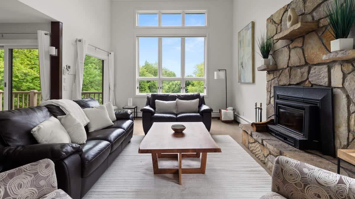Bright and airy living room featuring a large stone fireplace and high ceilings. Furnished with dark leather sofas, a wooden coffee table, and patterned armchairs, with large windows offering views of the surrounding nature.