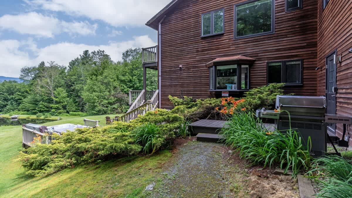 Exterior view of a rustic wooden house with multiple windows and a bay window, nestled amidst lush green landscaping and trees. A grill is visible on a small patio area, with a path leading to a hot tub and a grassy lawn in the background.