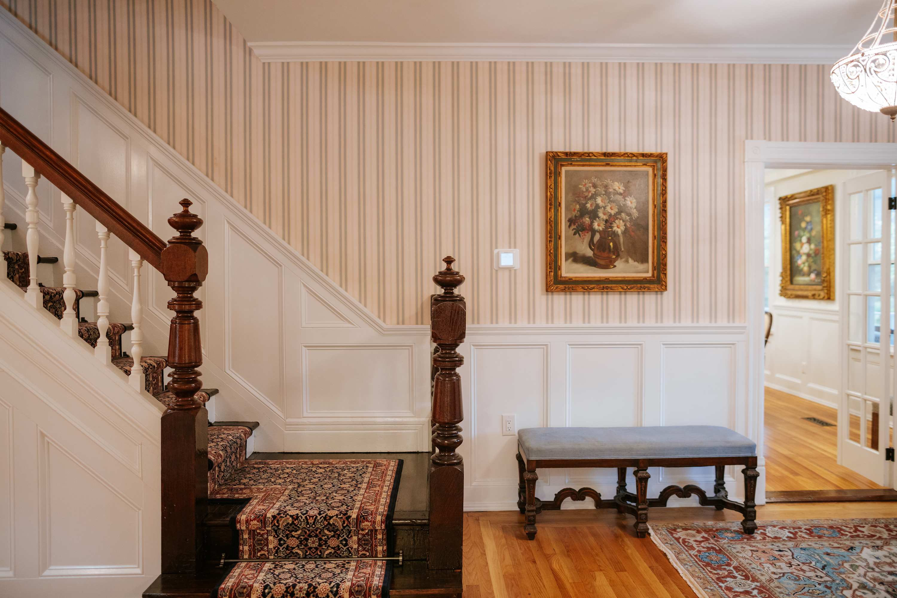 Grand foyer with a dark wood staircase featuring a patterned runner, white wainscoting, striped wallpaper, and hardwood floors, leading to another room through an open doorway.