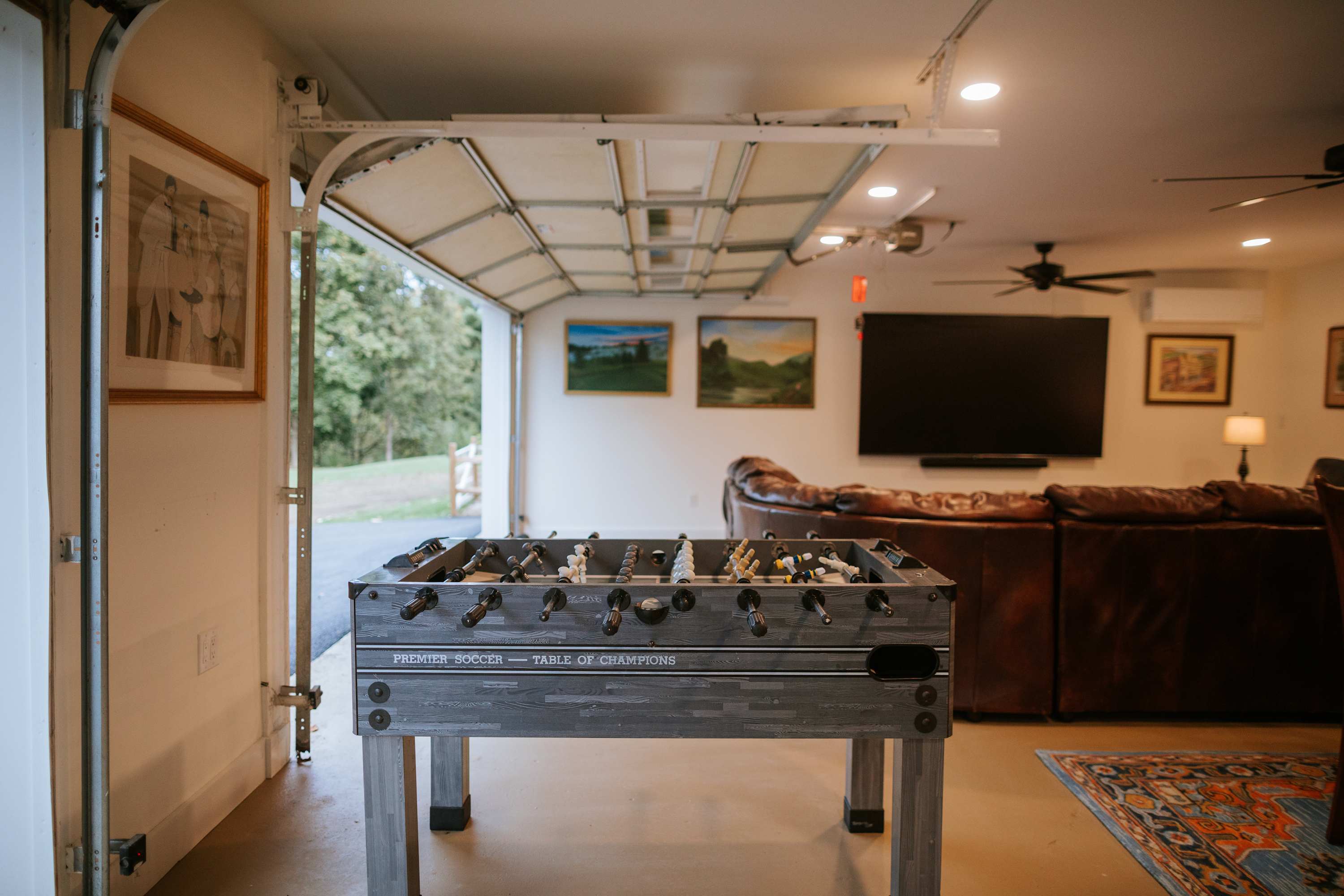 Game room with a foosball table in the foreground, a large brown leather sectional sofa, and a wall-mounted TV in the background. The space has a garage door that opens to the outside.