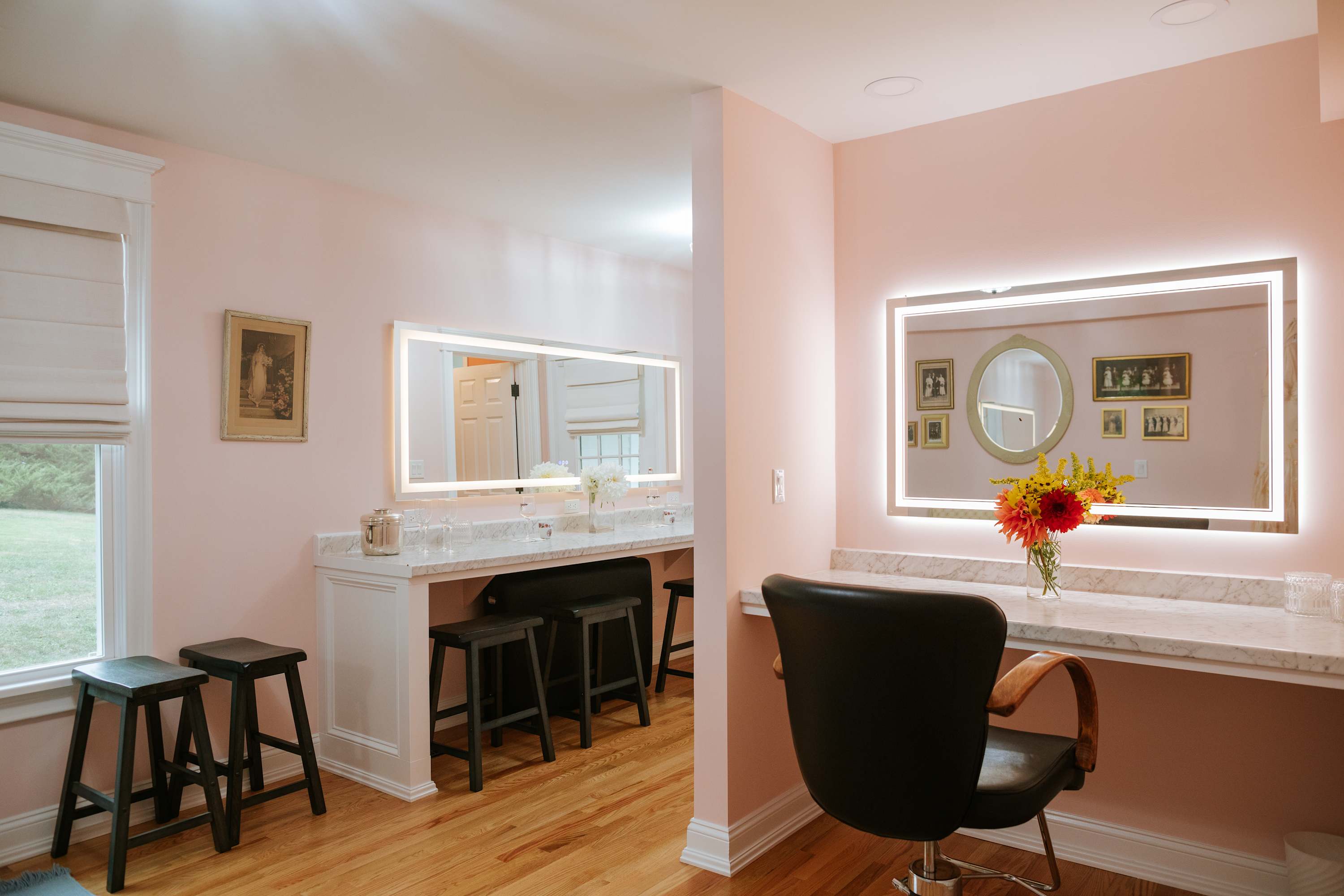 Bright vanity area with multiple lighted mirrors, marble countertops, and stools, set against light pink walls. A large window provides natural light, and a bouquet of flowers adds a decorative touch.