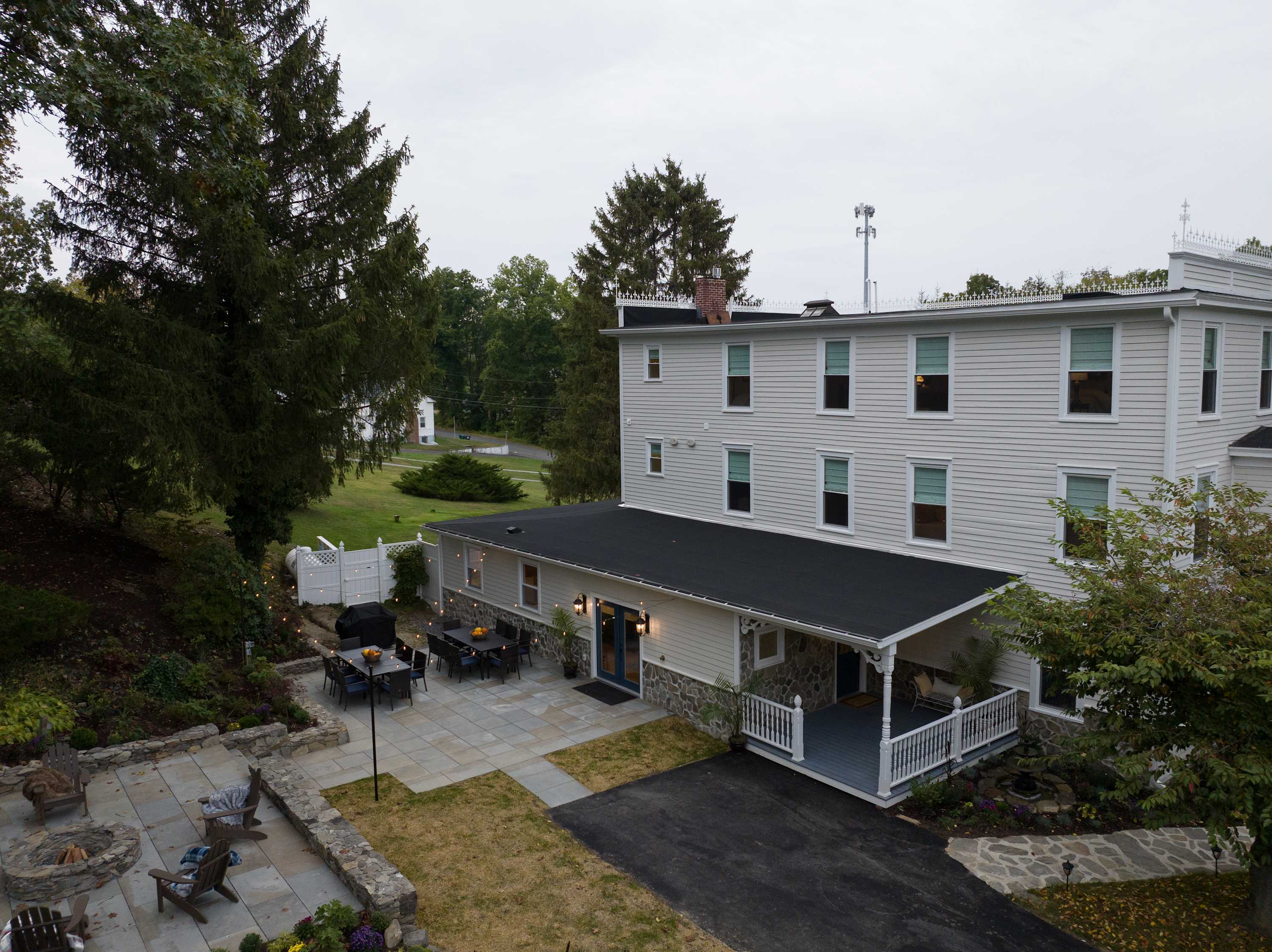The back exterior of the house featuring a stone patio with an outdoor dining area and a separate fire pit seating area. The house has white siding and a covered porch section, all surrounded by green lawn and trees.