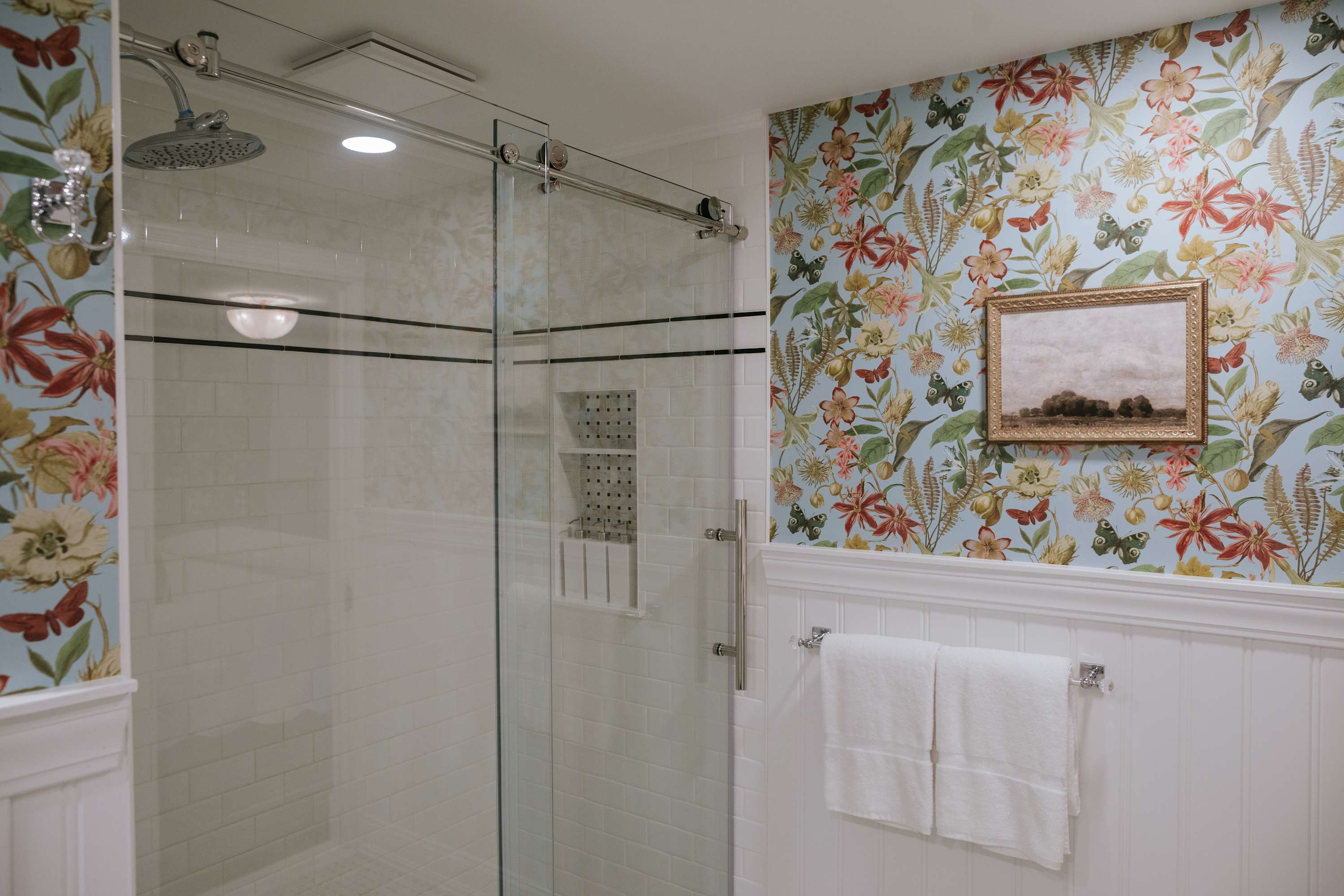 Bright bathroom featuring a spacious glass-enclosed shower with white subway tiles and a modern shower system. The walls are adorned with vibrant floral and butterfly wallpaper above classic white wainscoting.