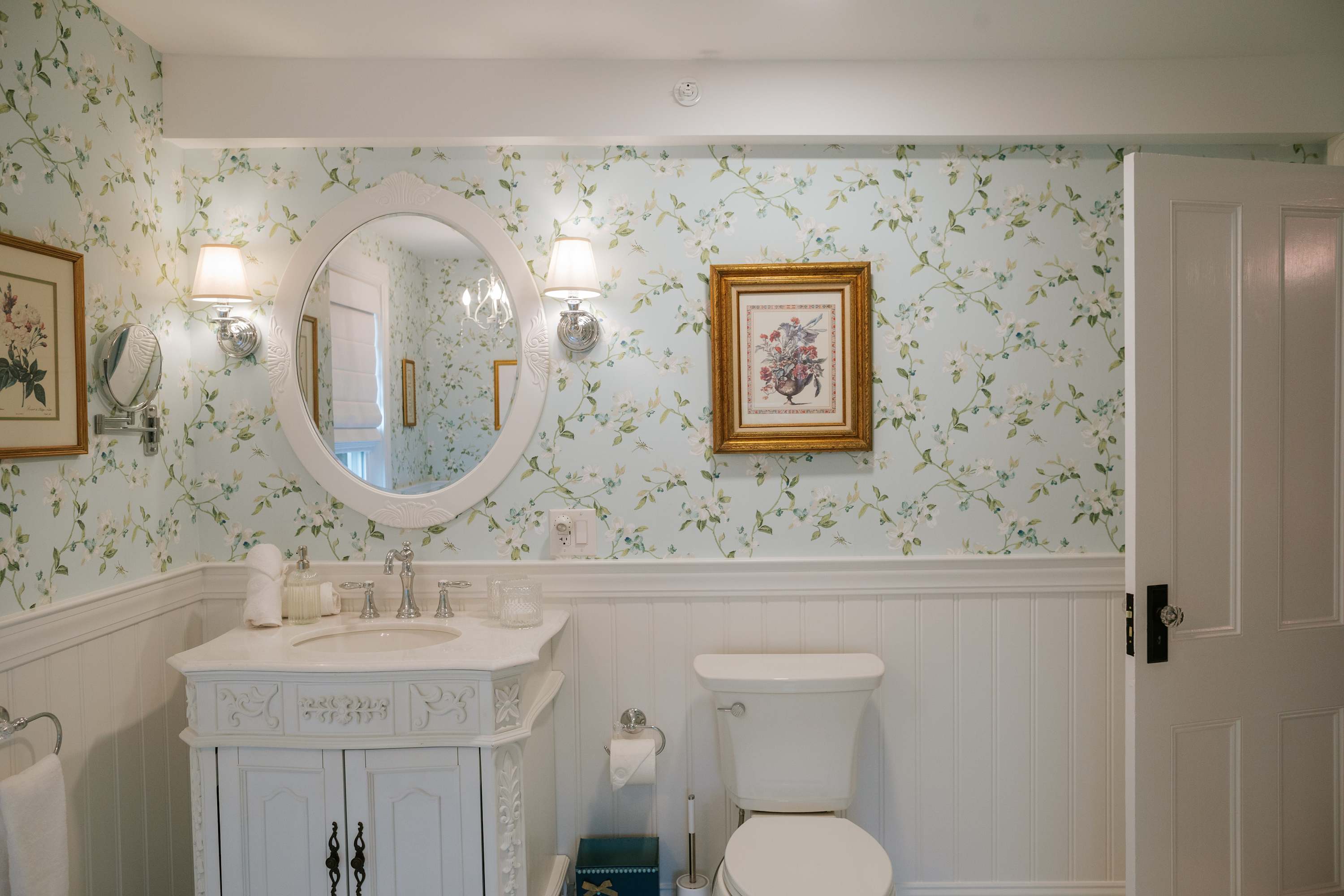 A charming bathroom with light blue floral wallpaper and white wainscoting, featuring a white vanity with a single sink, an oval mirror with sconce lighting, and a white toilet.
