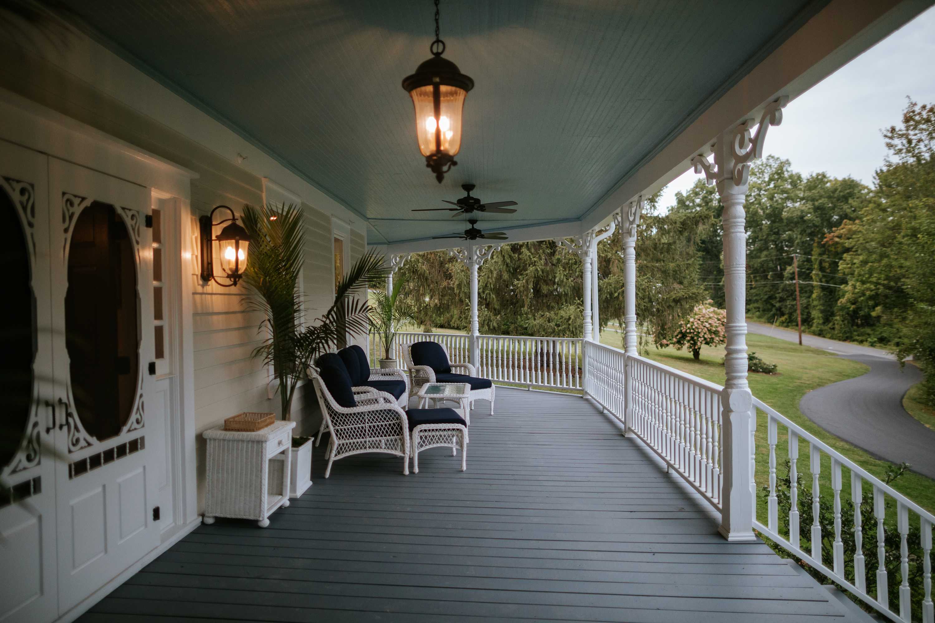 A view along the long wrap-around porch, featuring white wicker chairs with dark blue cushions and a light blue ceiling. Decorative white railings and columns line the porch, with hanging lanterns providing illumination.