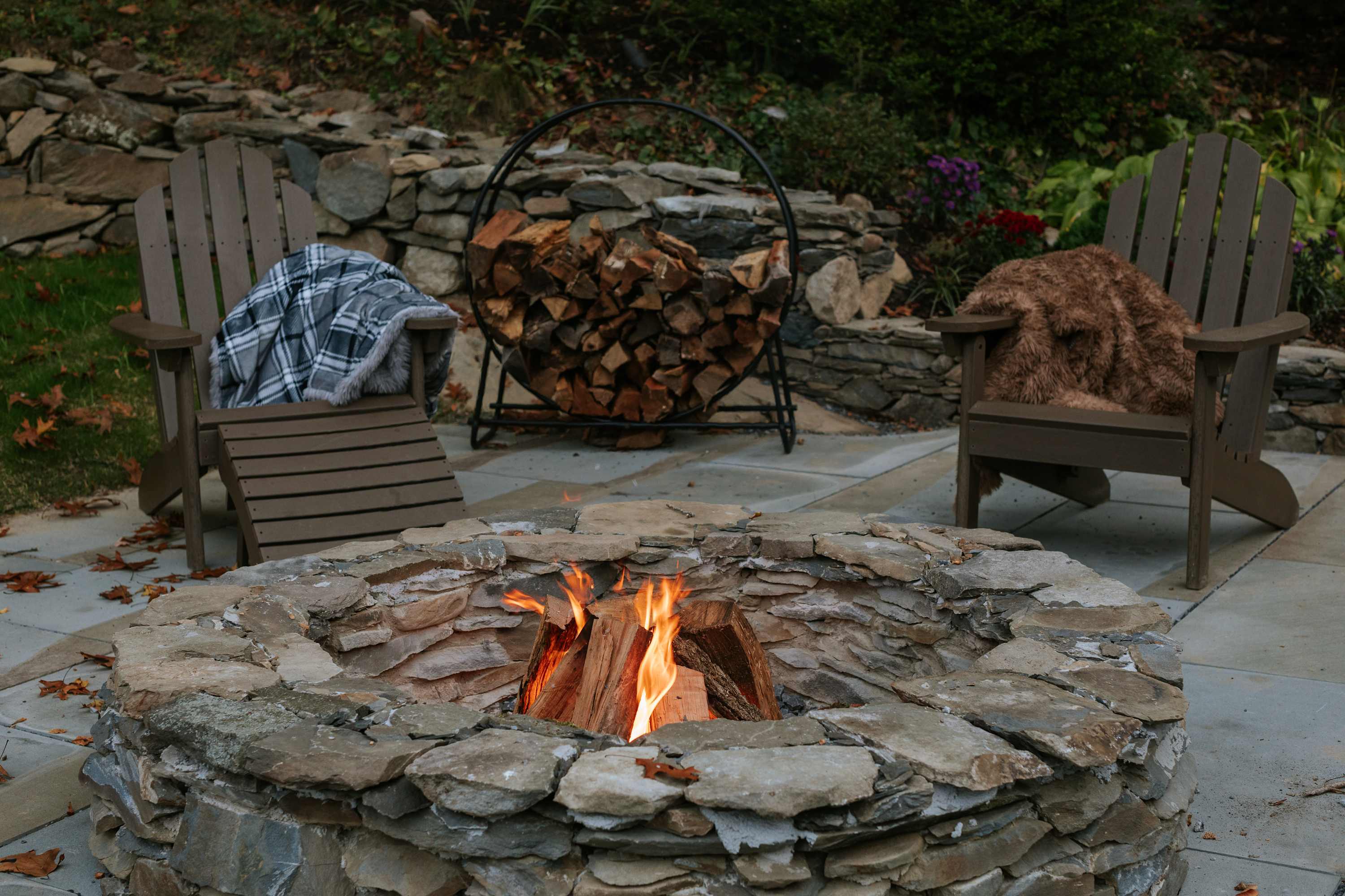 Close-up view of a rustic stone fire pit with burning logs, surrounded by two wooden Adirondack chairs with blankets. A metal log holder filled with firewood is visible in the background.