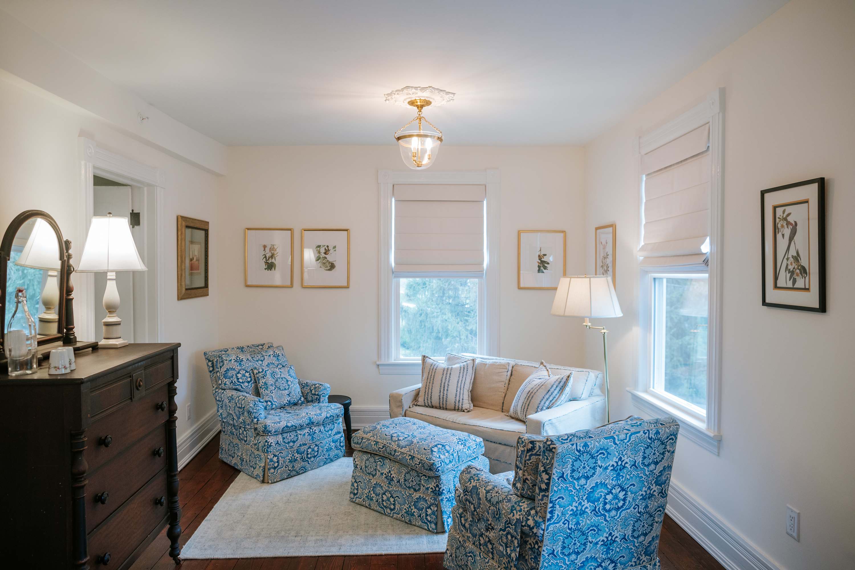 A bright sitting area within a bedroom, featuring a cream sofa and two blue patterned armchairs with a matching ottoman, arranged on a light rug. Dark hardwood floors, multiple windows with Roman shades, and a dark wood dresser are visible.