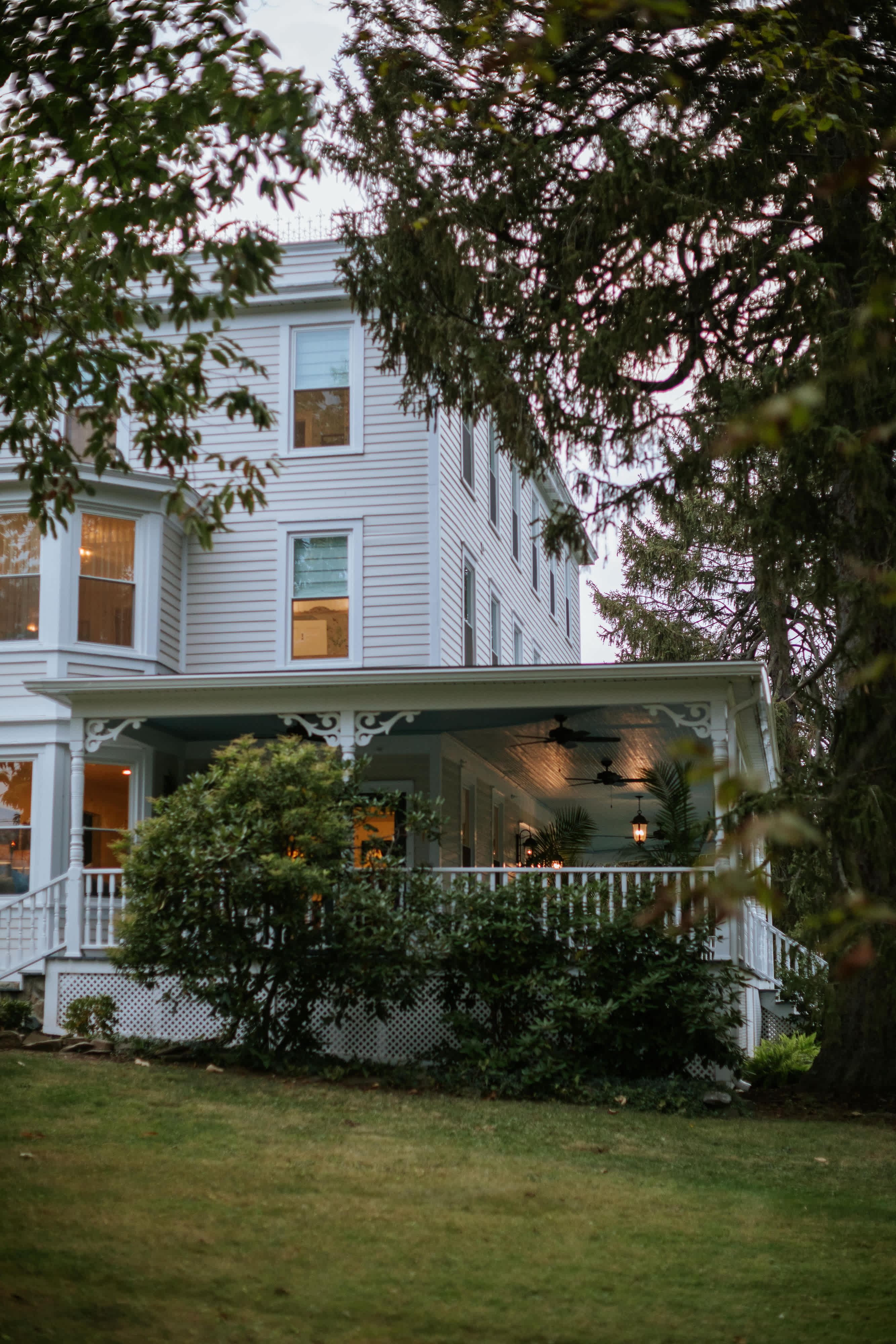 A grand white clapboard house showcases a wide, covered porch with intricate white railings and a ceiling fan, nestled among mature trees and green foliage. The porch offers a welcoming entrance with visible interior lighting.