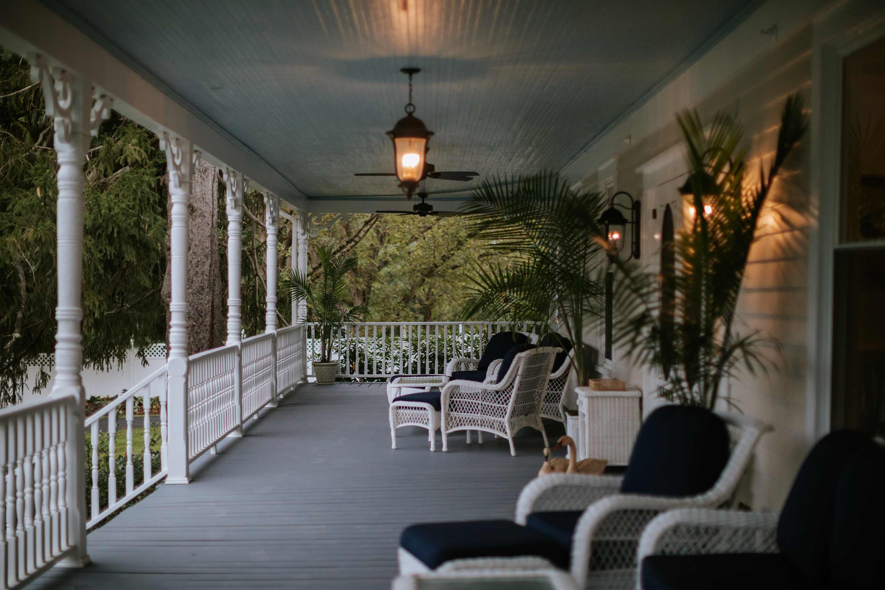 Inviting wrap-around porch with light blue ceiling, white wicker furniture with dark cushions, and potted plants. Overhead lights and ceiling fans provide comfort, overlooking lush greenery.