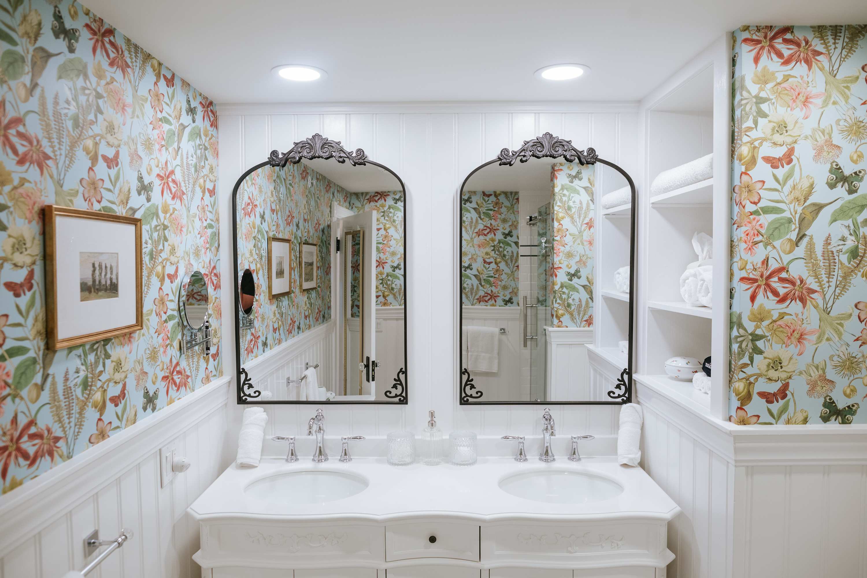 A bright bathroom with a double white vanity featuring two sinks and chrome faucets, flanked by two arched mirrors with ornate black frames, set against light blue floral wallpaper and white wainscoting.