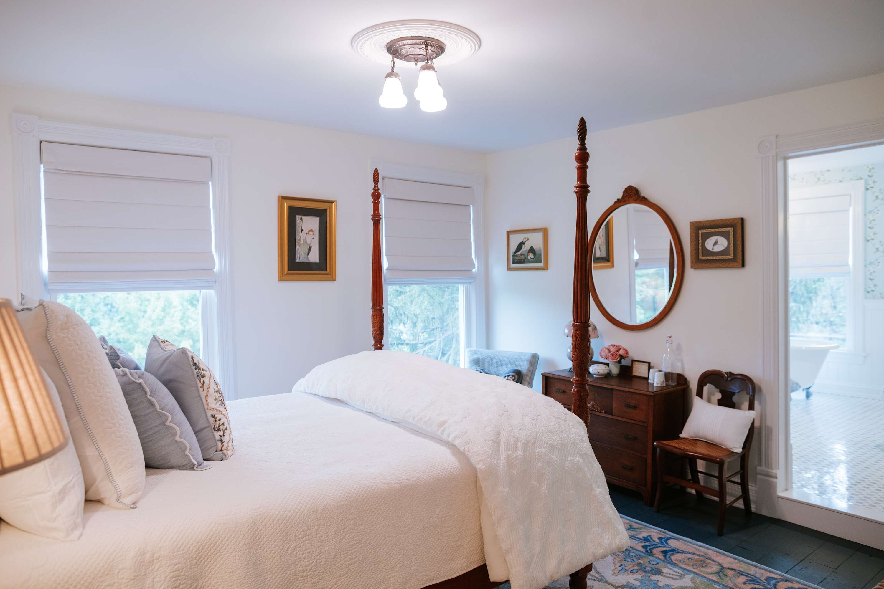 An elegant bedroom with a dark wood four-poster bed dressed in white bedding, a matching wooden dresser with an oval mirror, and a patterned area rug, with a glimpse of a floral wallpapered bathroom through an open doorway.