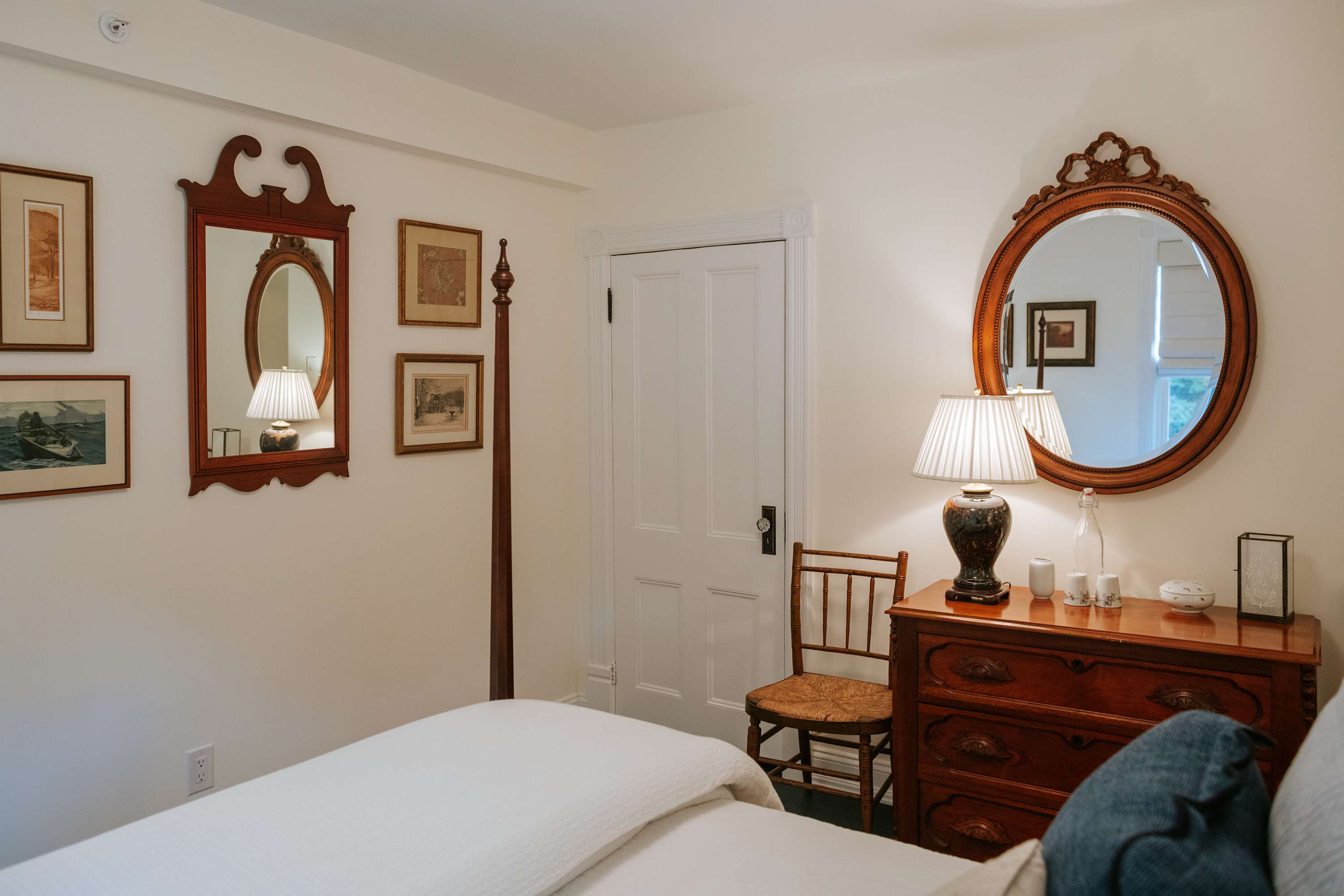 A cozy bedroom featuring a bed with white bedding, a dark wood dresser with an ornate mirror above it, and a bedside lamp with a pleated shade, all against white walls with framed artwork.