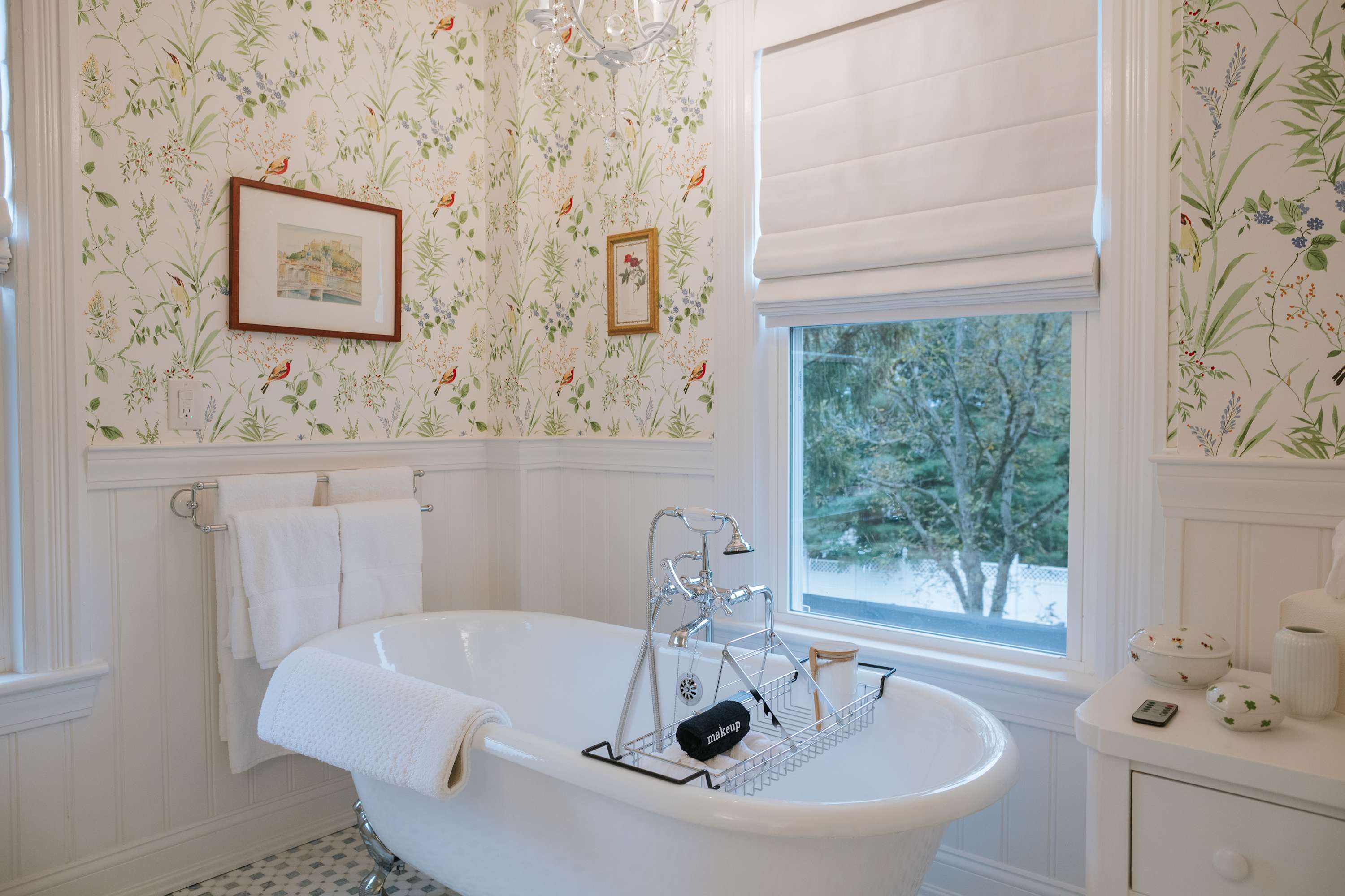 A charming bathroom with a freestanding clawfoot tub and a large window covered by a white Roman shade. The walls feature white wainscoting and floral wallpaper, with a small framed picture and a towel rack nearby.
