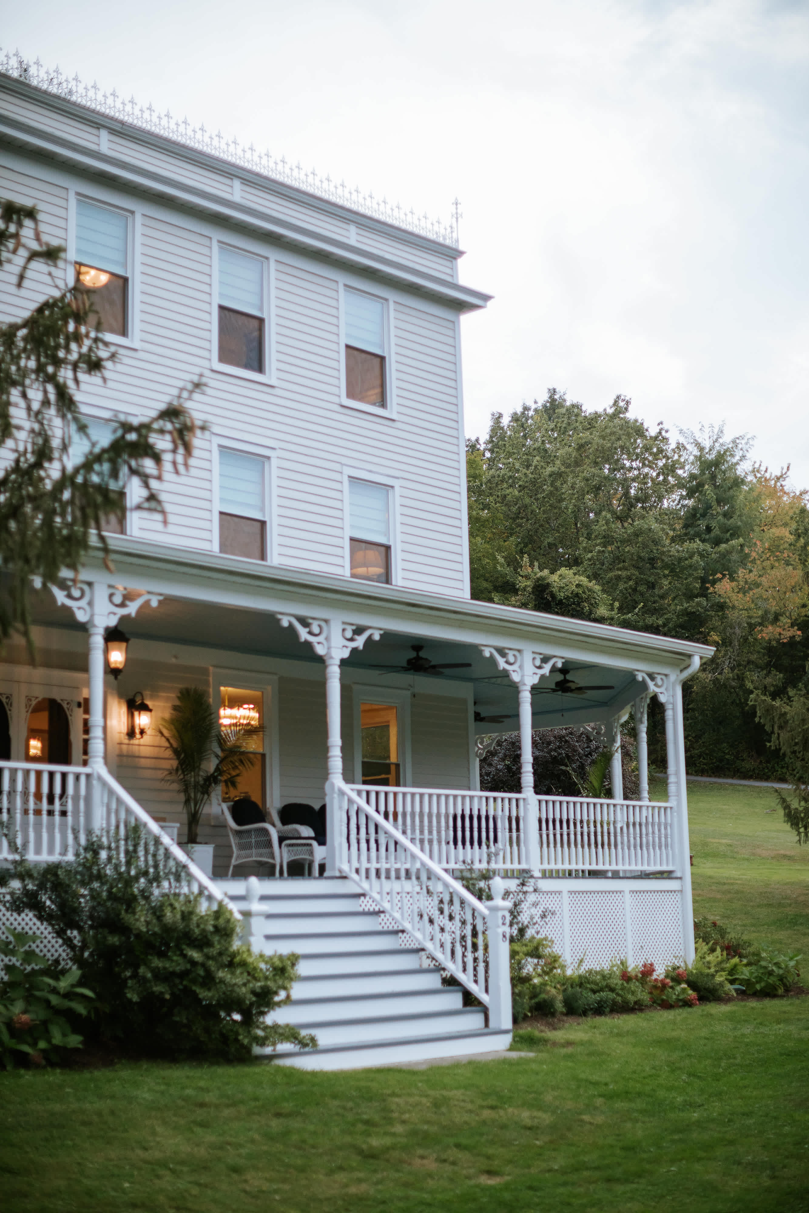 Side view of the large white multi-story house, showcasing the wrap-around porch with white railings and steps. Lush green lawn and trees surround the property under an overcast sky.