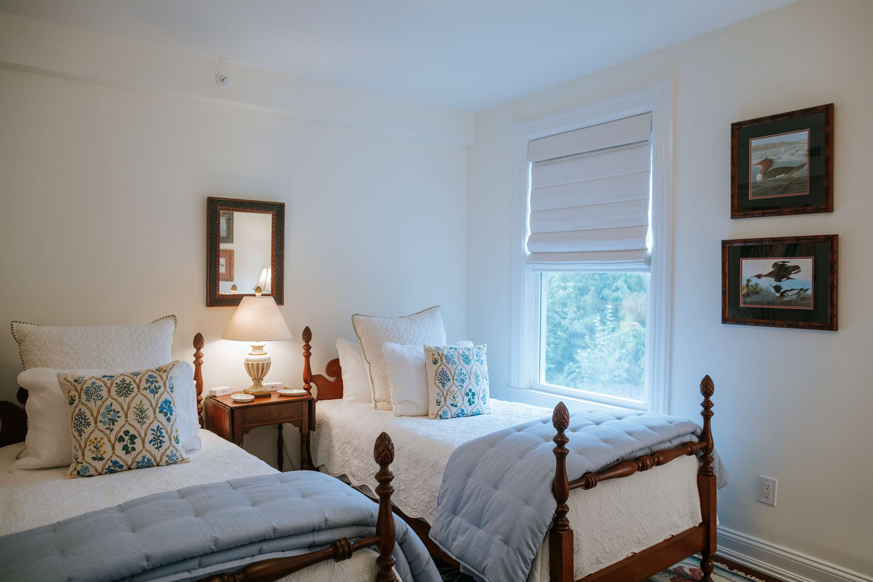 A bright bedroom with two twin beds featuring dark wood frames and white quilted bedding with blue accents, separated by a small wooden nightstand with a lamp. White walls and a window with a Roman shade are visible.