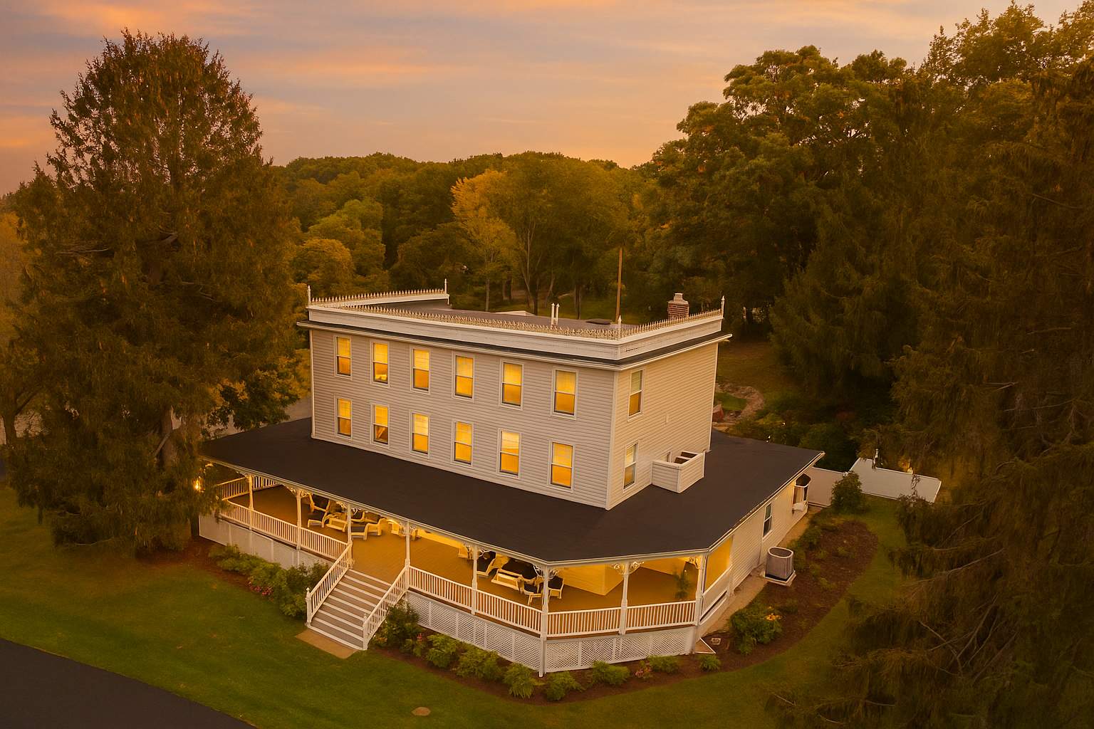 Aerial view of a large, light-colored multi-story house with a wide wrap-around porch, set amidst lush green trees under a dramatic sunset sky. The house features multiple windows and a decorative roofline.