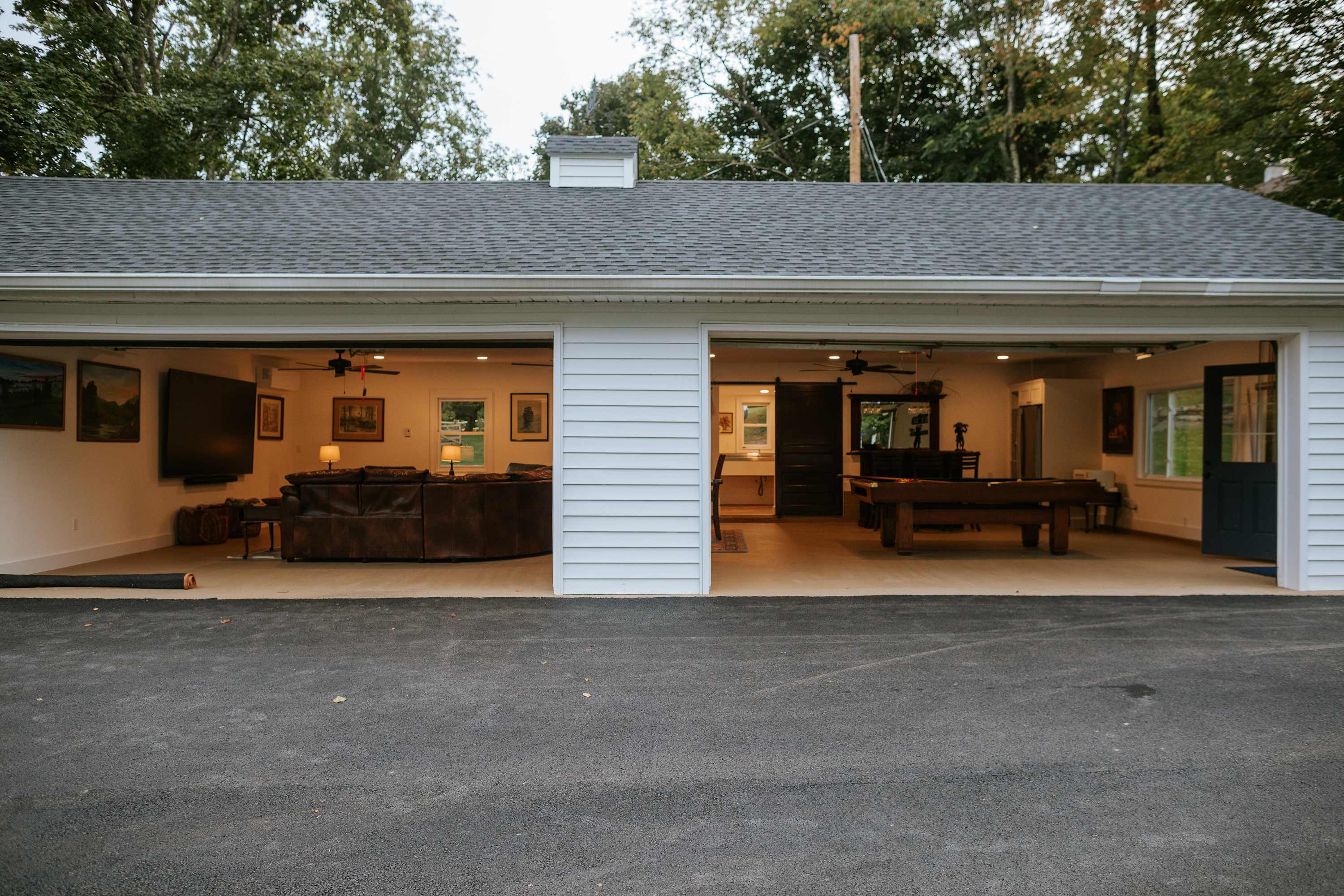 The exterior of a white, two-bay garage conversion is shown with both doors open, revealing a cozy living area with a large sofa on the left and a game room with a pool table on the right. The asphalt driveway leads up to the structure, surrounded by trees.