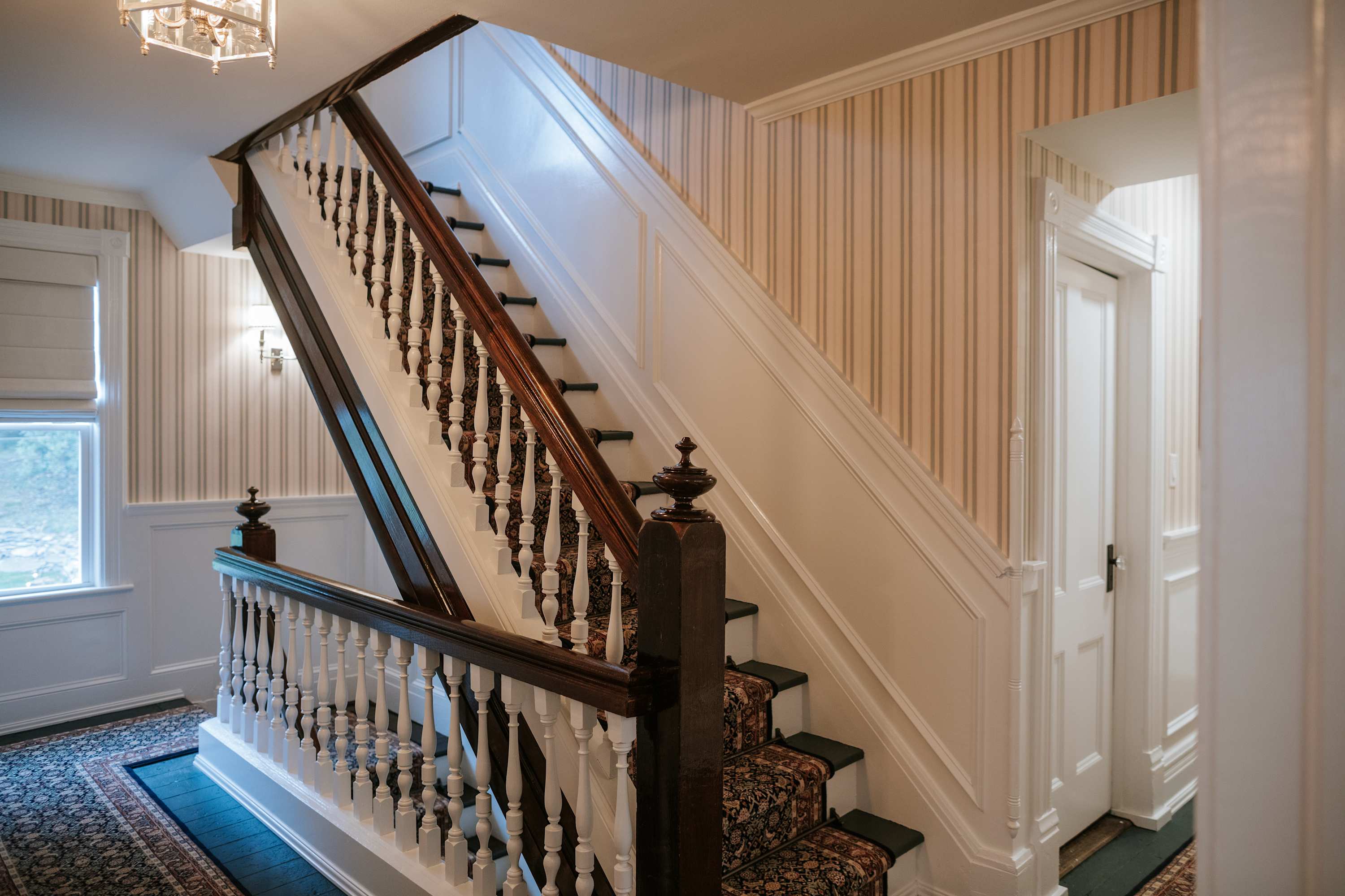 Elegant staircase featuring dark wood handrails, white balusters, and a patterned carpet runner, set against walls with white wainscoting and classic striped wallpaper. A chandelier hangs above, illuminating the space.