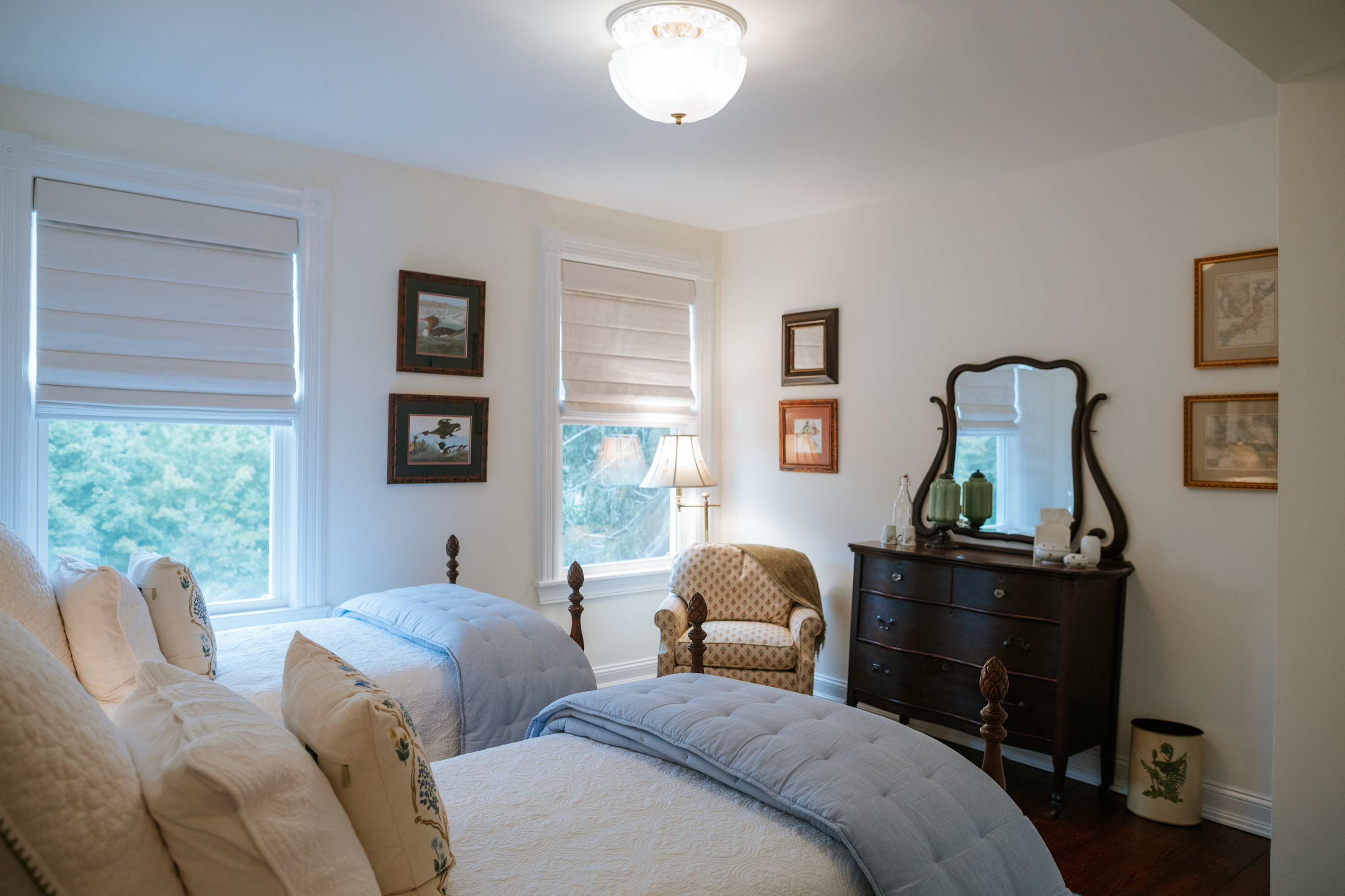 A cozy bedroom with two twin beds featuring dark wood frames and white quilted bedding, a dark wood dresser with an antique mirror, and a patterned armchair. White walls and two windows with Roman shades are present.