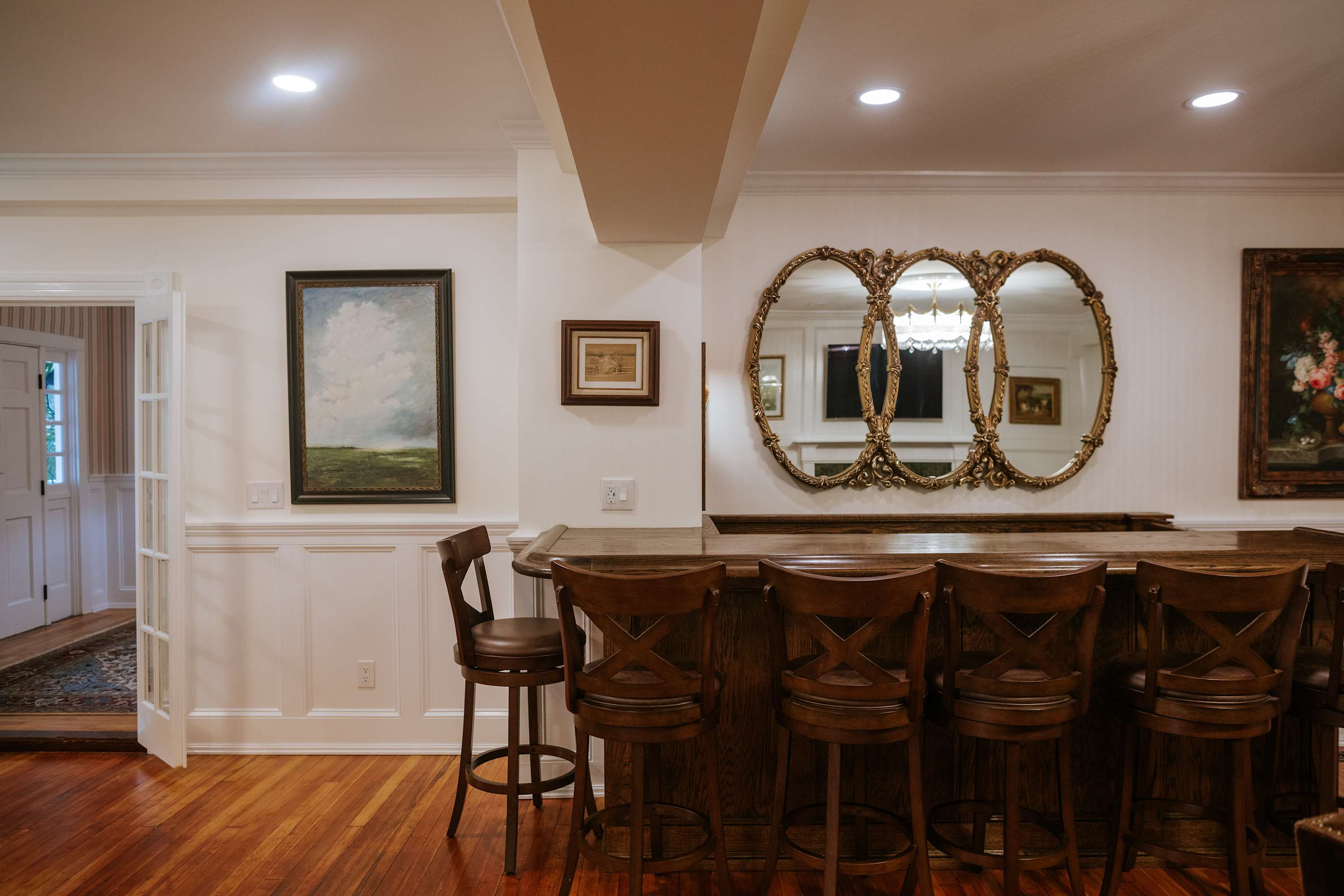 Entertainment area featuring a wooden bar with stools, white wainscoting, and hardwood floors. Decorative mirrors and artwork adorn the walls, creating a classic and inviting atmosphere.