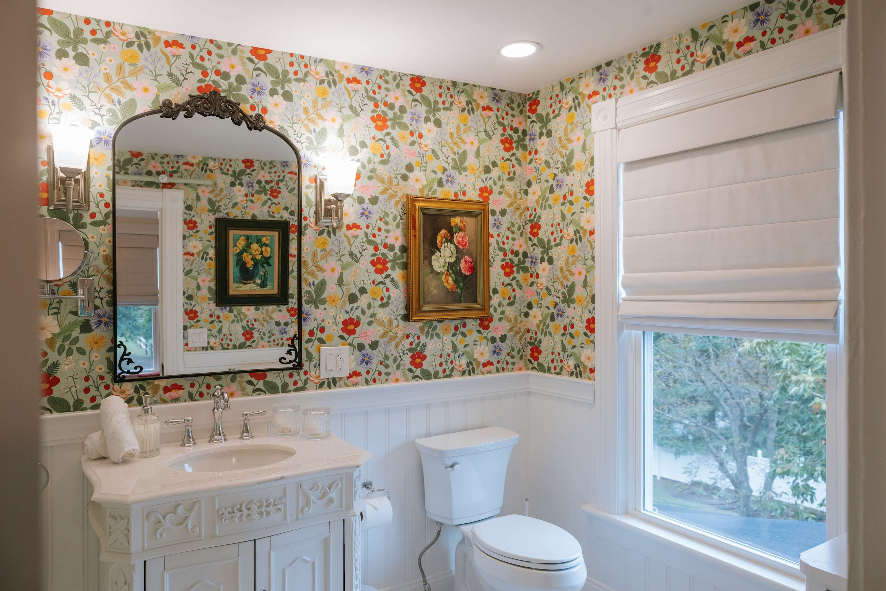 A vibrant bathroom with floral wallpaper featuring red, yellow, and green flowers, white wainscoting, a white vanity with a marble top, and an ornate black mirror. A window with a white Roman shade is visible.