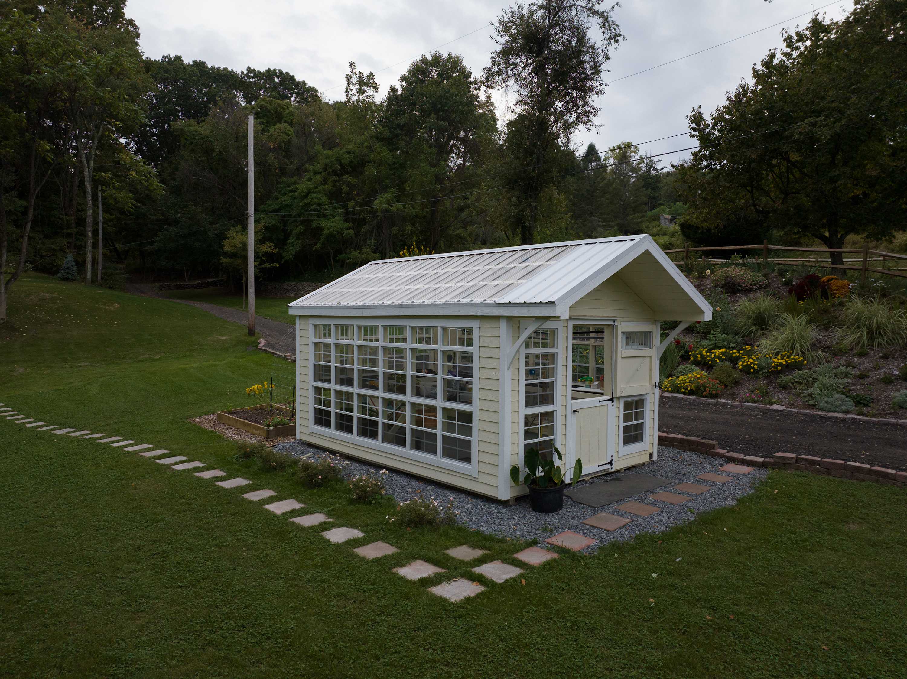 A charming yellow greenhouse with numerous windows and a white roof, situated in a lush green garden. Stepping stones lead up to the entrance, and various plants and flowers are visible around it.