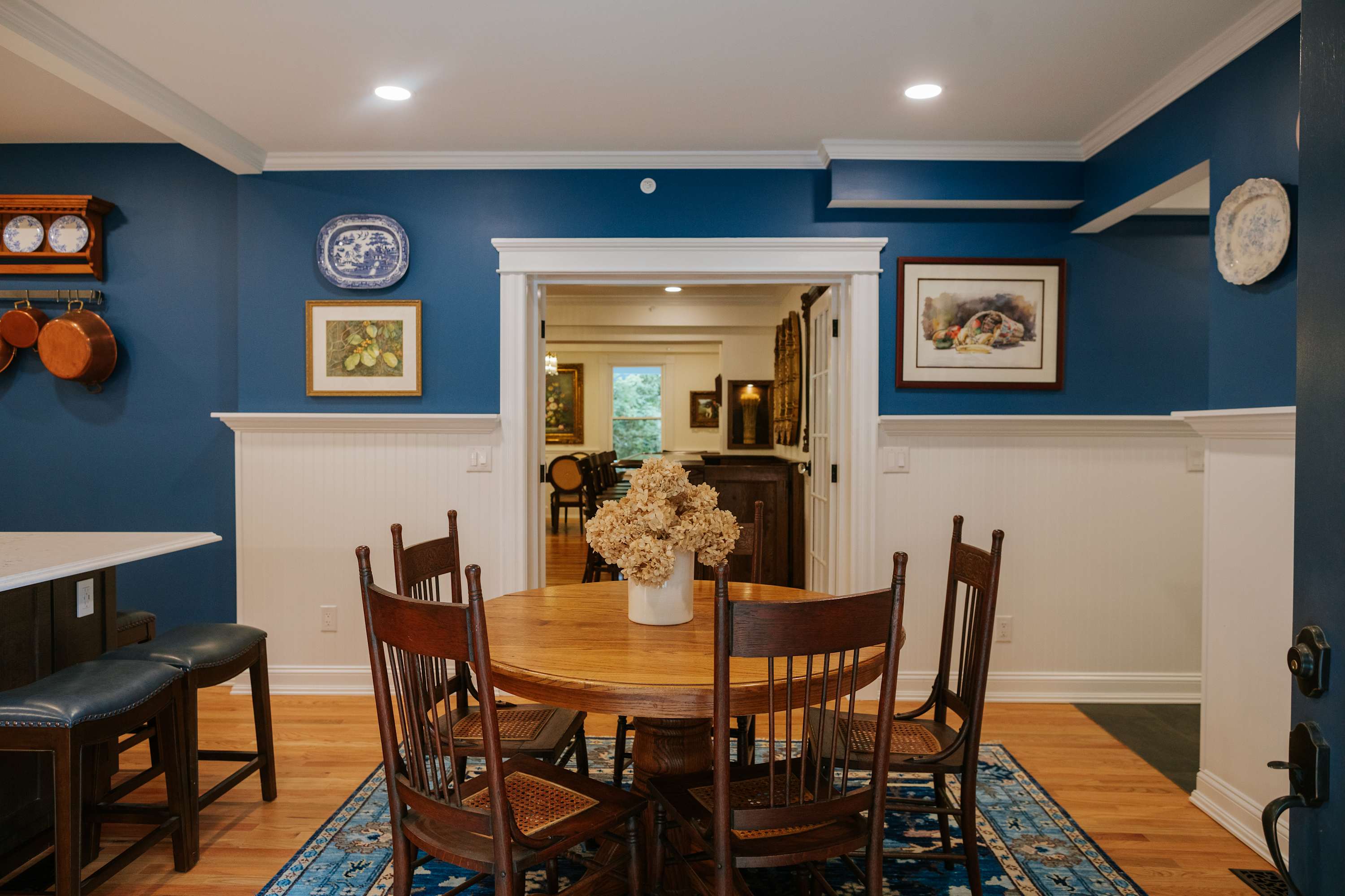 Cozy kitchen dining nook with a round wooden table and four chairs on a blue patterned rug, featuring blue walls with white wainscoting and an open doorway to another room.