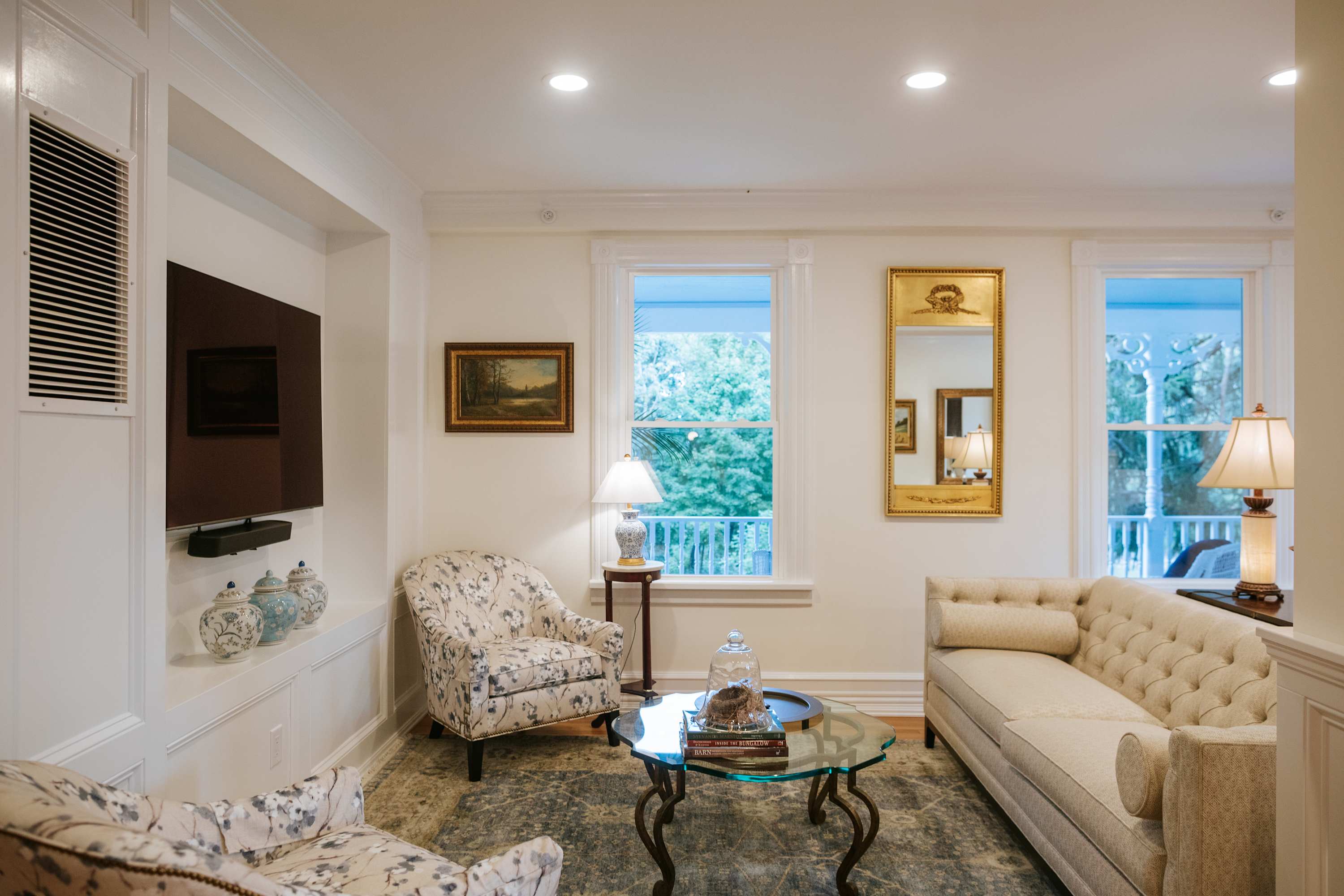 Bright living room with white walls, a tufted beige sofa, a floral armchair, a glass coffee table, and a wall-mounted TV, featuring large windows overlooking greenery.