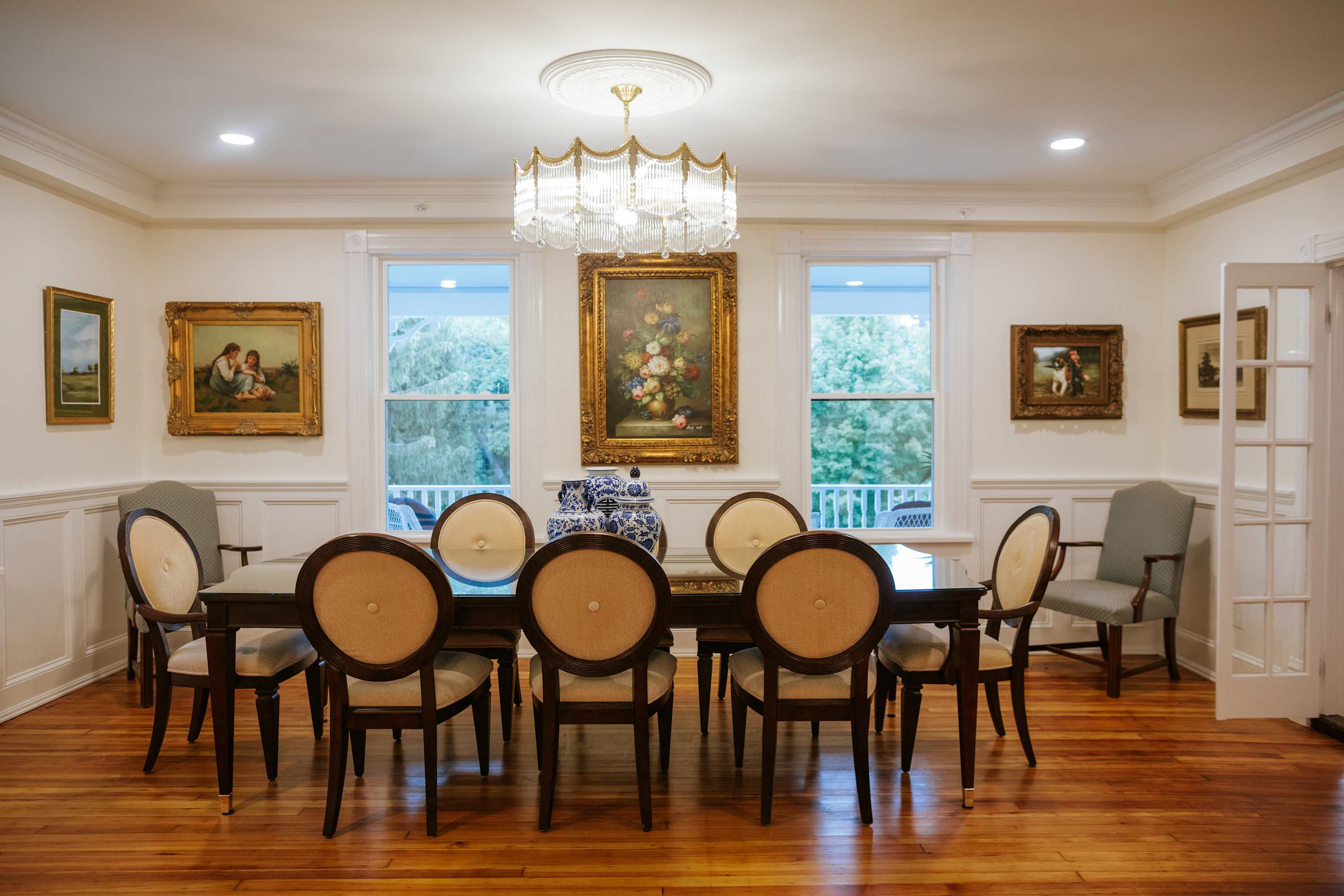 Formal dining room with a long dark wood table, eight upholstered chairs, a crystal chandelier, and multiple windows, adorned with classic artwork and a large decorative vase.