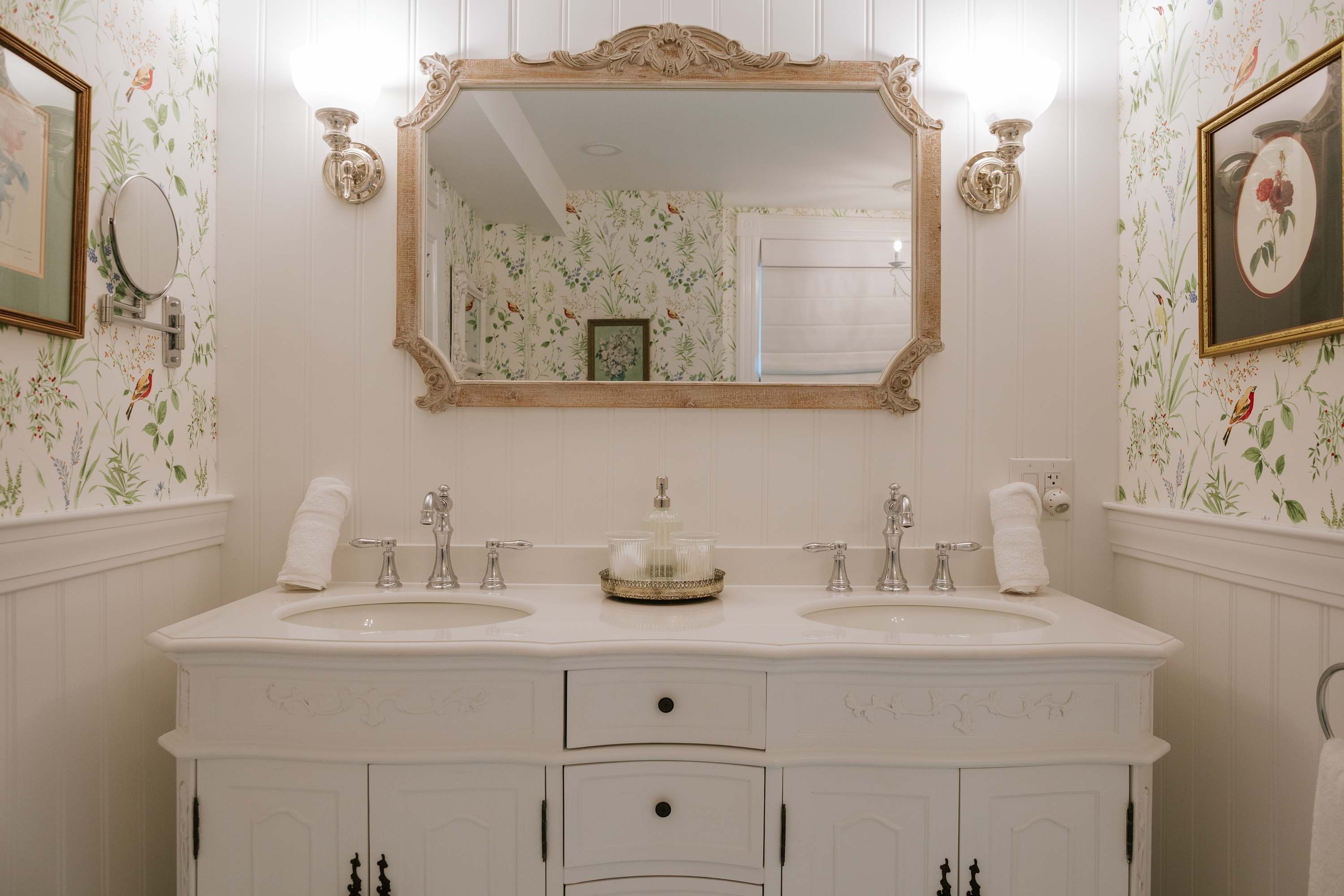 A bright bathroom with a double vanity featuring white cabinets, two sinks, and chrome faucets. The walls are adorned with white wainscoting and floral wallpaper with birds, complemented by an ornate mirror and sconce lighting.