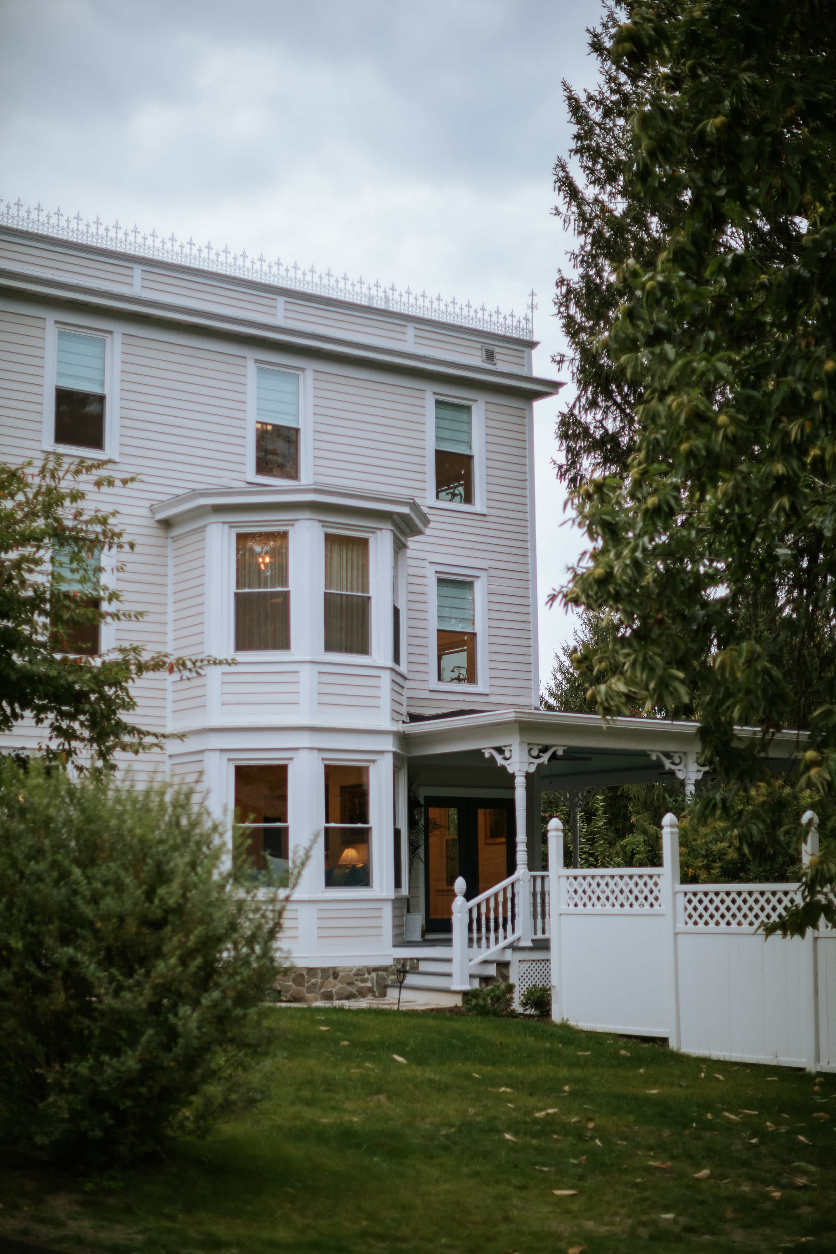 A charming multi-story white clapboard house features an ornate bay window on the left and a covered porch with white railings and decorative trim on the right. Lush green landscaping and mature trees frame the elegant Victorian-style architecture.