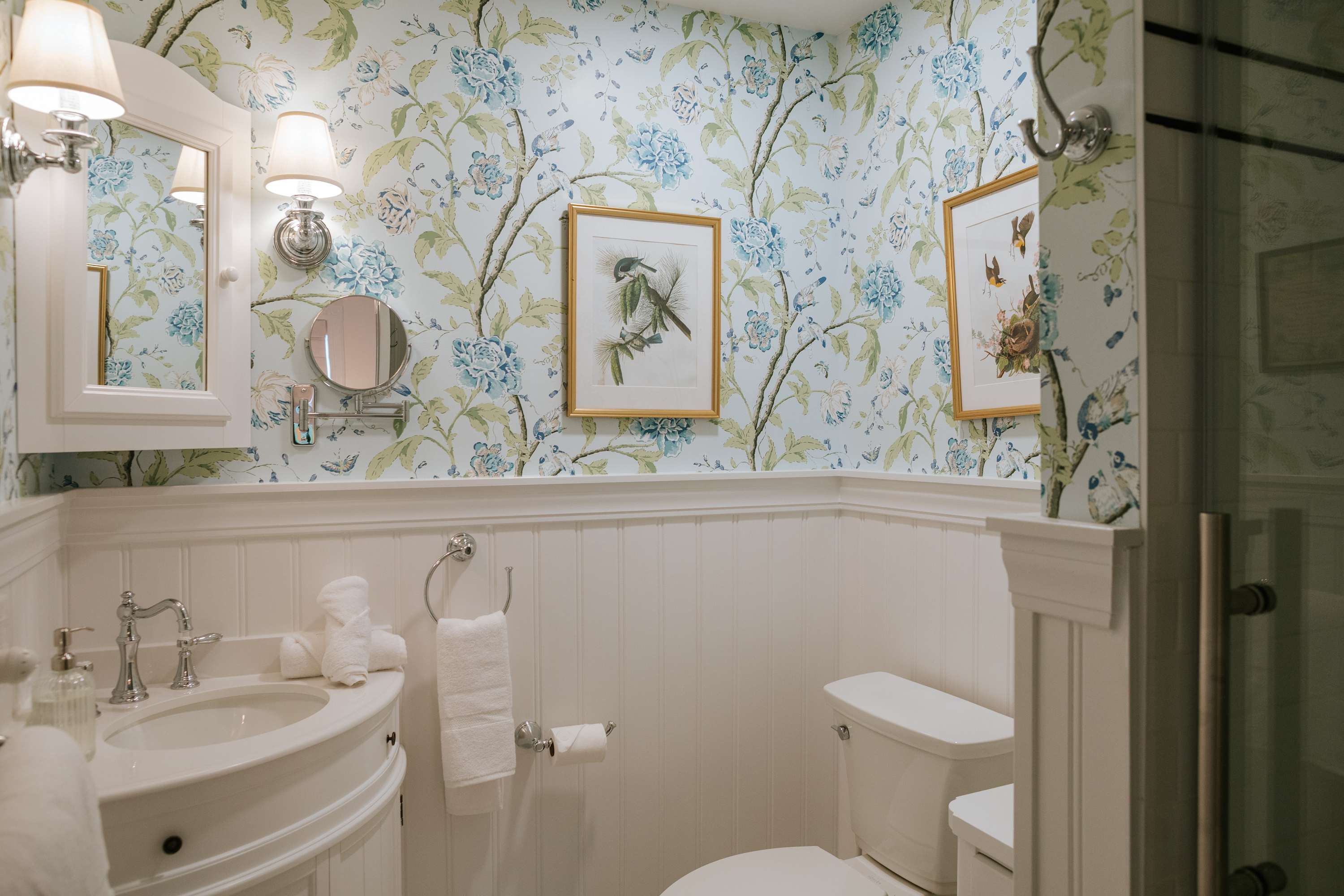 A charming bathroom with blue and green floral wallpaper, white wainscoting, a white pedestal sink, and a toilet. A glass shower enclosure is partially visible, along with a wall-mounted mirror and sconces.